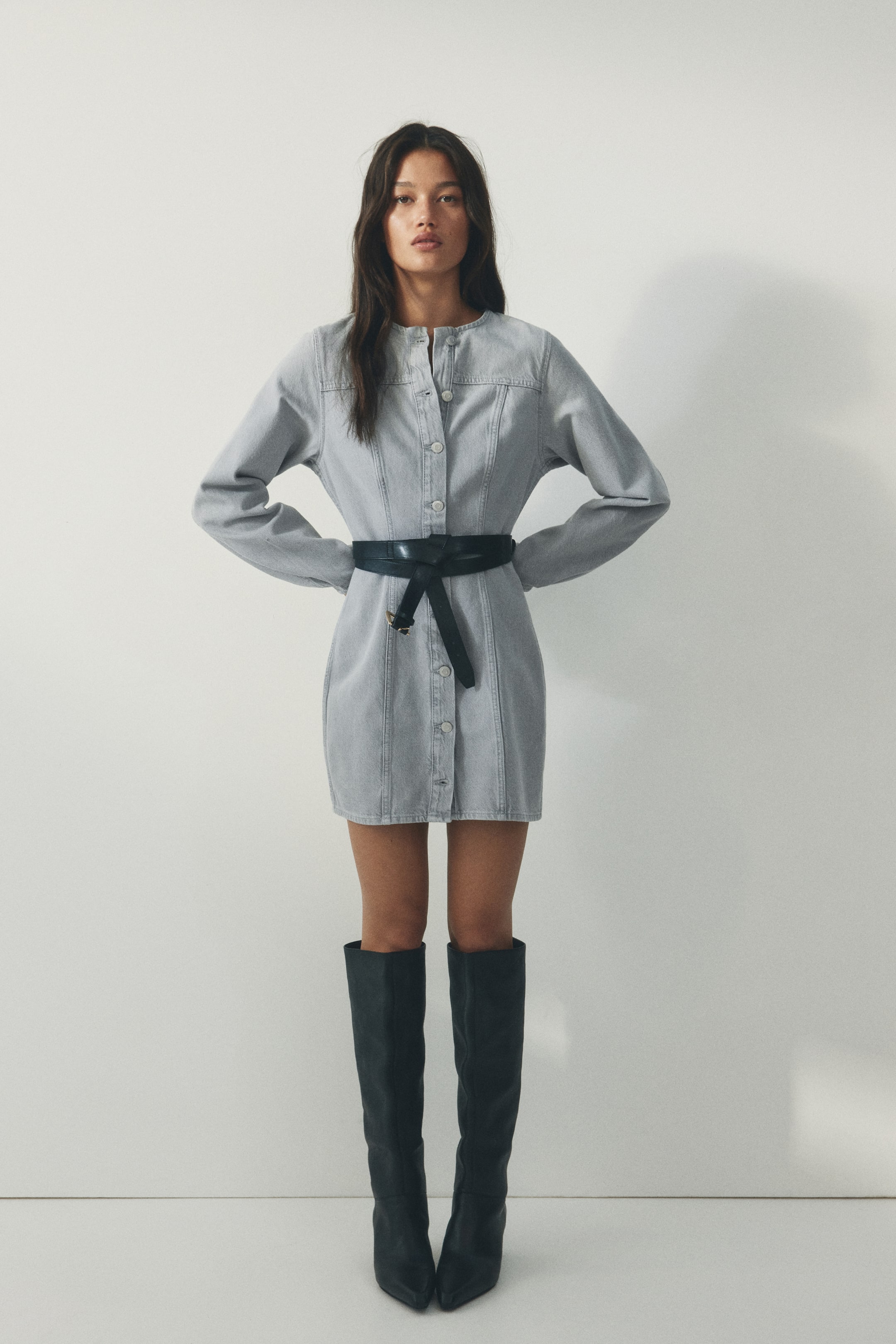 Größeres Bild ansehen: Ein hellgraues Denim-Kleid mit Stehkragen, langen Ärmeln und einer durchgehenden Knopfleiste mit vertikalen Nahtdetails. Ein schwarzer Bindegürtel betont die Taille des Kleides.