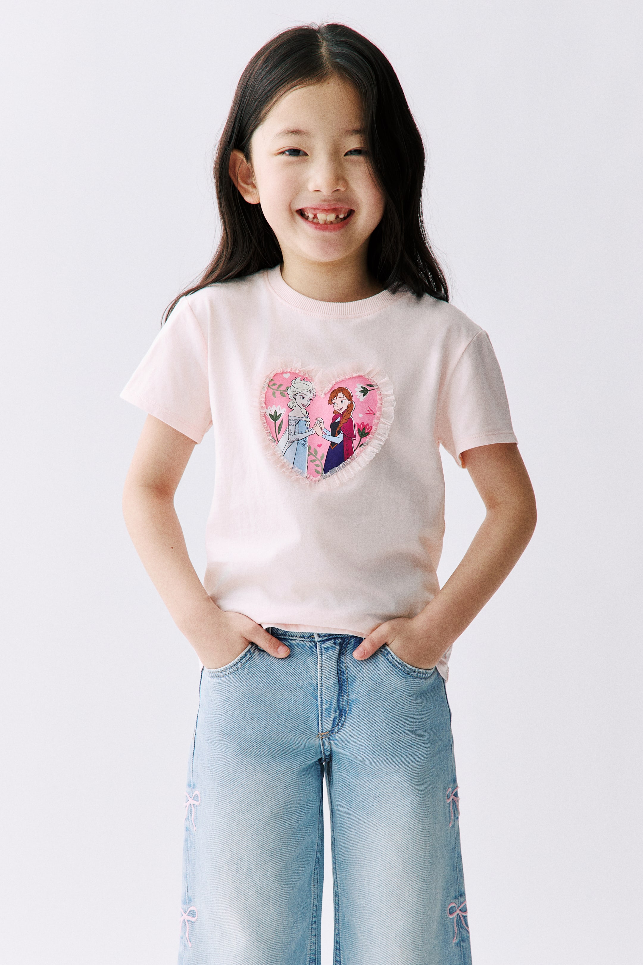 拡大画像を表示: モデルが着用しているのは、ライトピンクのTシャツで、柔らかいプリントコットンジャージー素材にリブネックラインが施されています。フロントには、2人のアニメ風女性キャラクターのハート型グラフィックが描かれており、フリルのトリムで縁取られています。