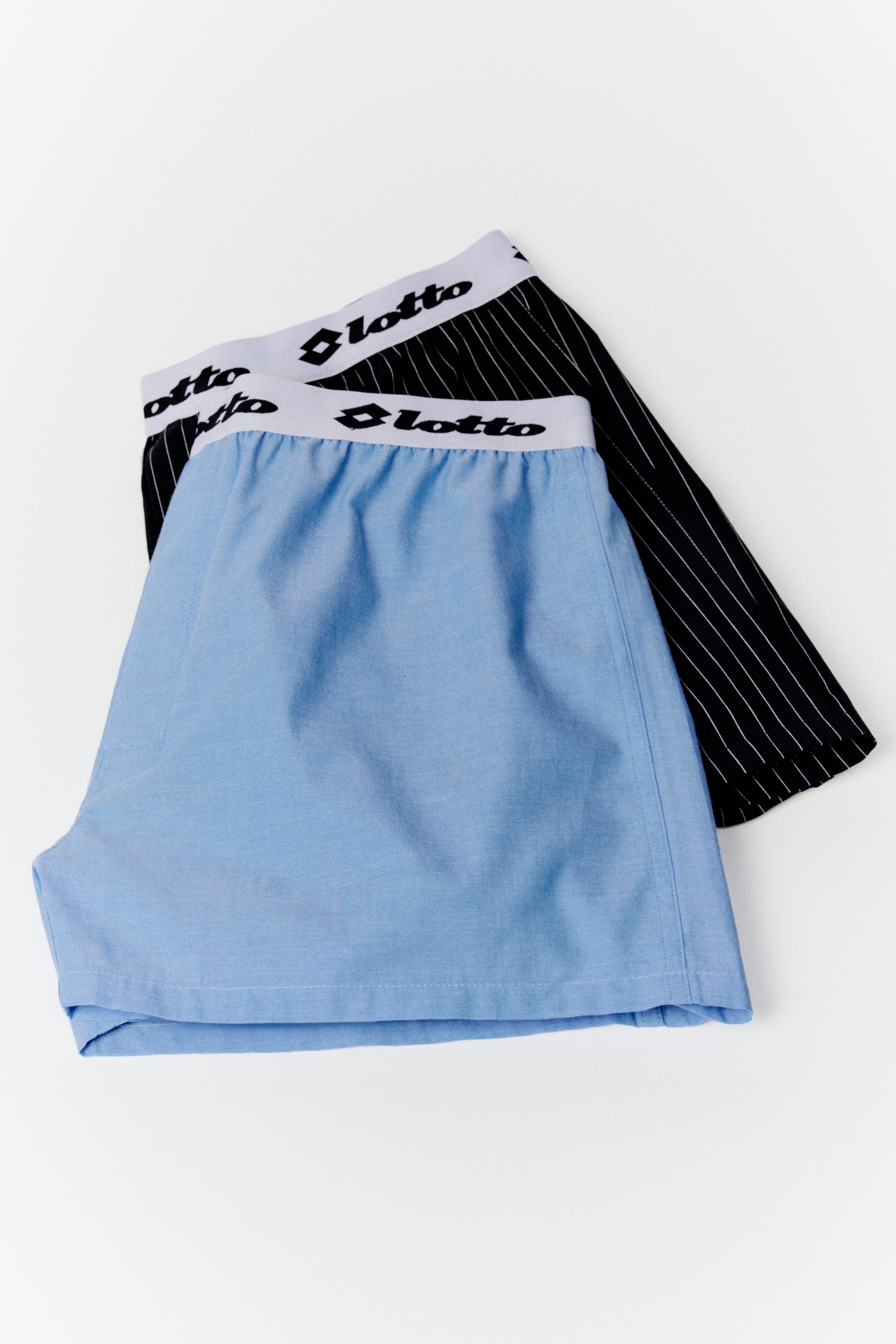 View larger image: Two men's boxer shorts are showcased, with a light blue pair draped partially over a black pinstriped pair. Both boxers feature white elastic waistbands adorned with the black "Lotto" logo.