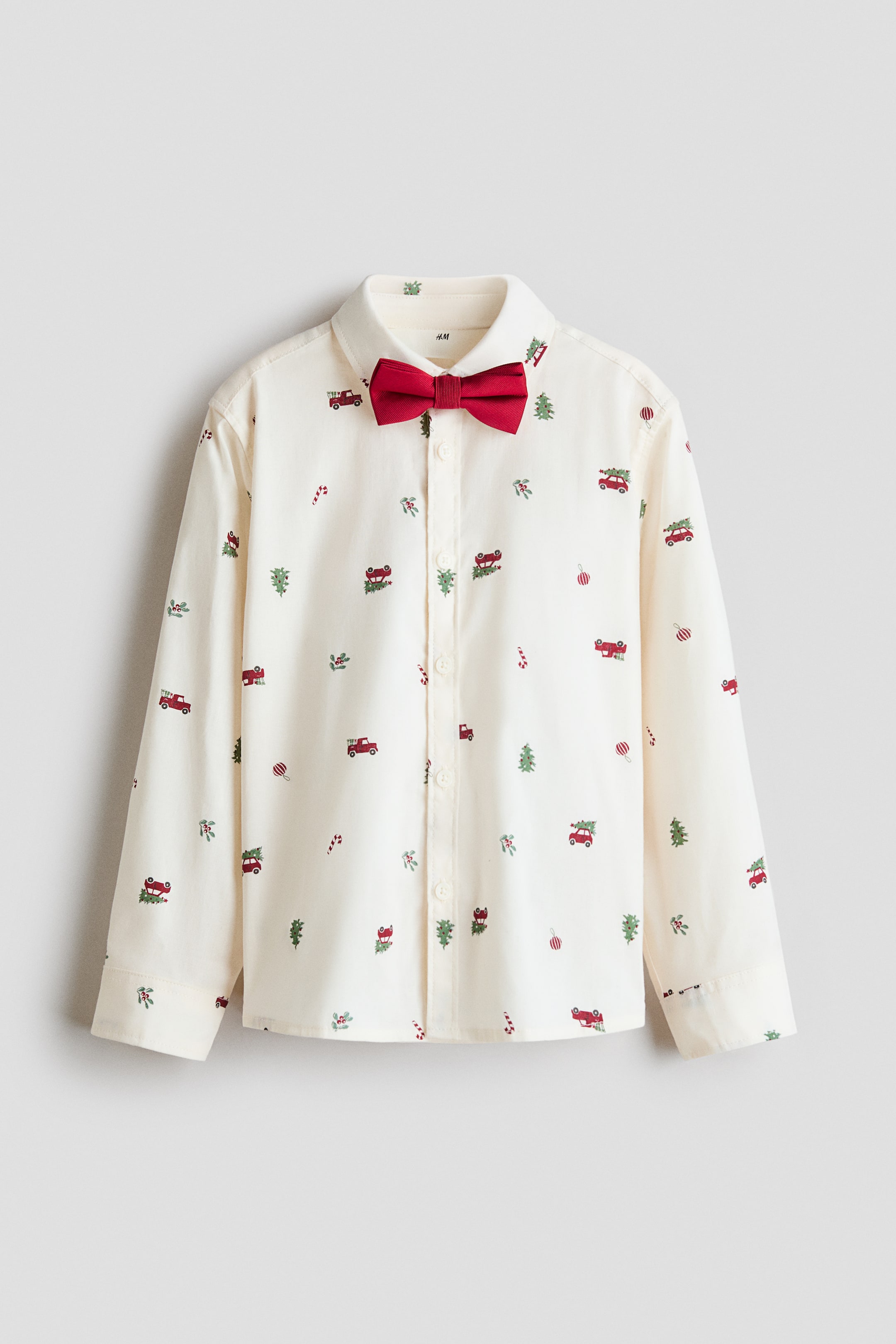 View larger image: A long-sleeved, cream-colored shirt features a holiday-themed pattern of small red trucks, green trees, candy canes, and ornaments. The shirt is styled front-facing with a solid red pre-tied bow tie at the turn-down collar and includes a button-down front with buttoned cuffs.