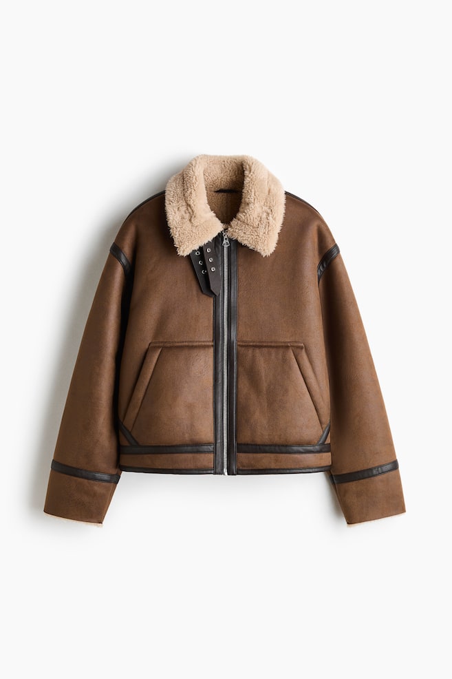 En brun aviatorjakke i shearling er vist forfra med en markant cremefarvet faux-shearling-krave og en gennemgÃ¥ende lynlÃ¥s foran. Designet fremhÃ¦ver mÃ¸rkebrune faux-lÃ¦derkanter langs sÃ¸mmene, lynlÃ¥sen, nederkanten og manchetterne, samt skrÃ¥ paspolerede forlommer og dobbelte halsrem-detaljer ved kraven. - 1