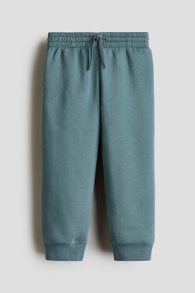 Un par de pantalones de chándal azul verdoso cuelga visto de frente, mostrando una cintura elástica con cordón y discretos bolsillos laterales. Los pantalones se estrechan ligeramente hacia la parte inferior y terminan en puños anchos acanalados. - 1