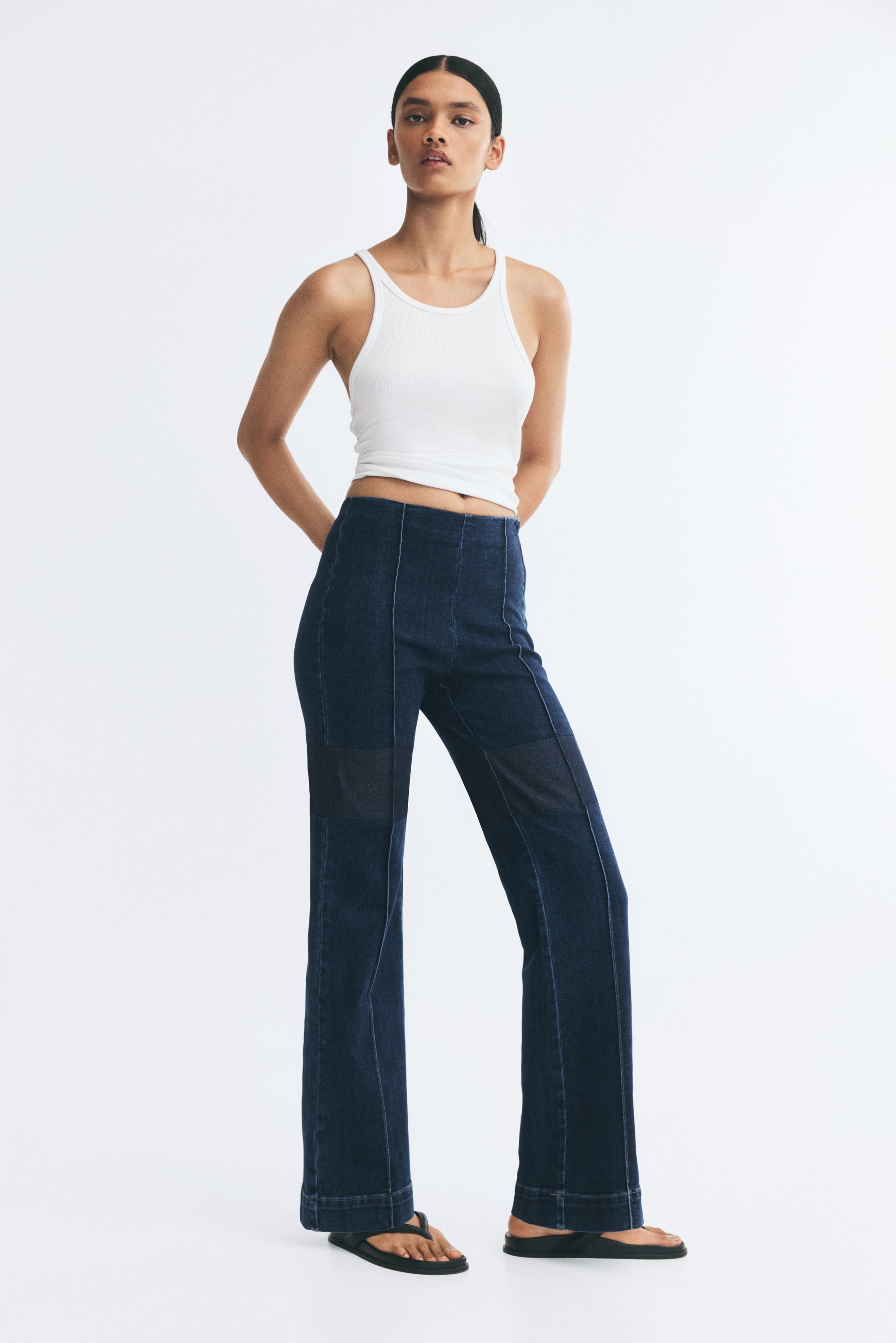 A pair of dark wash denim trousers is displayed from the front, featuring a high-rise waist and a wide-leg silhouette. Vertical stitched seams run down the centre of each leg, ending at wide, double-stitched hems.