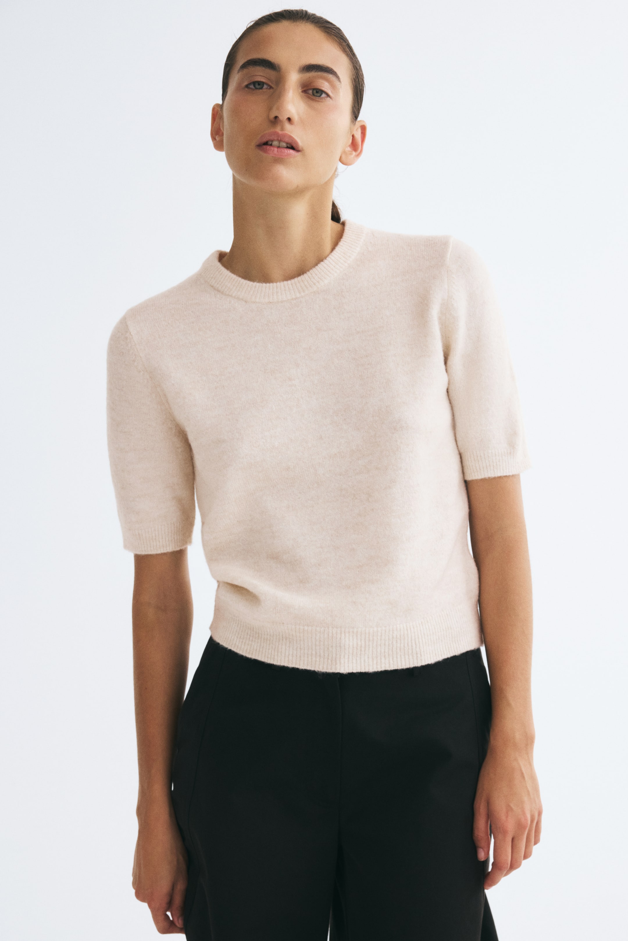 Ver imagen más grande: La modelo lleva puesta una camiseta de manga corta de punto suave, en beige claro, con cuello redondo acanalado, puños acanalados y bajo recto acanalado. La camiseta se combina con pantalones negros.