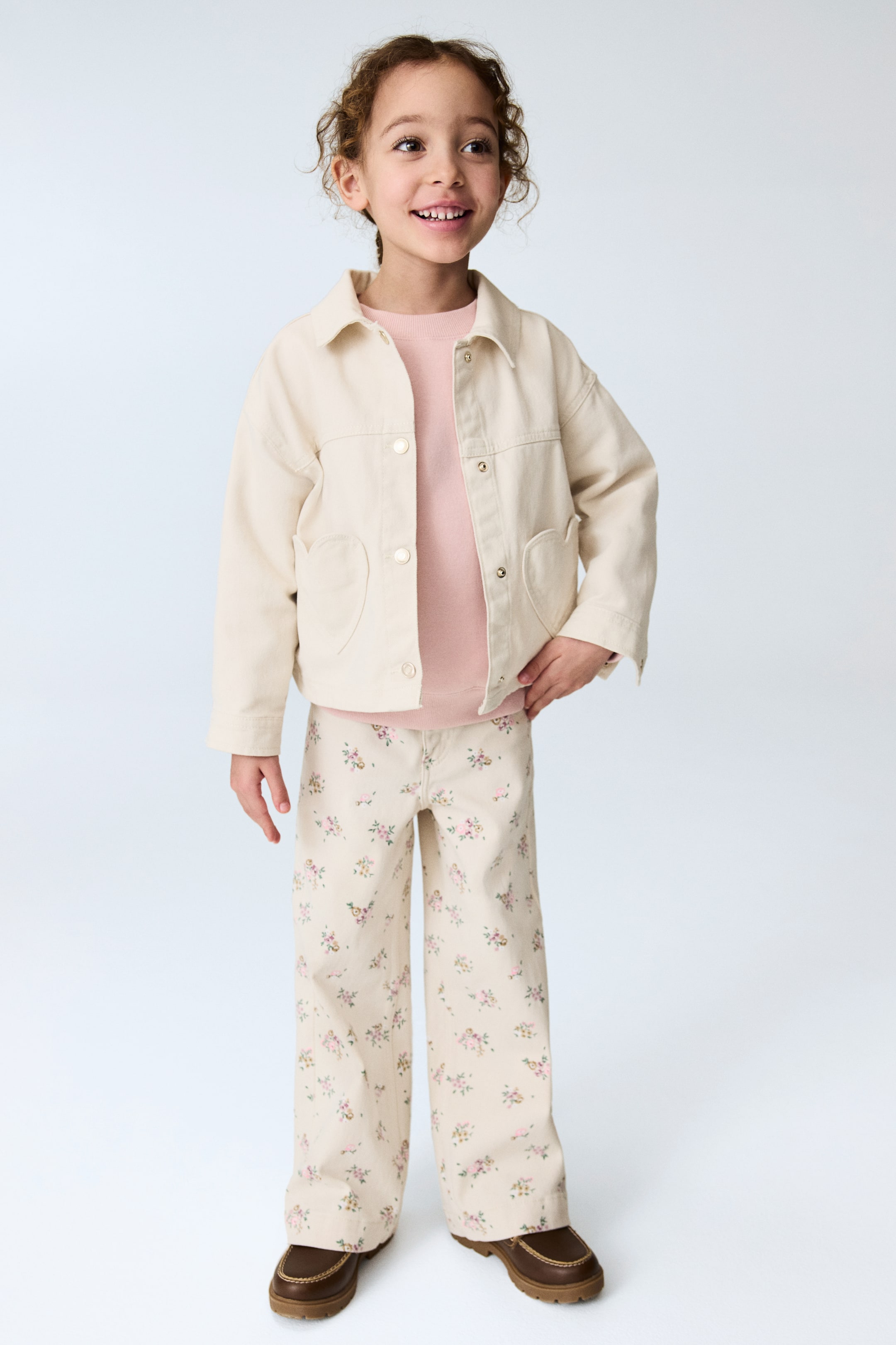 View larger image: A cream utility jacket with a classic collar, front snap button closure, and two rounded patch pockets is styled open over a light pink crew neck jumper. Coordinating wide-leg trousers in cream with an all-over small floral print are paired with dark brown moccasin-style shoes featuring a chunky lug sole.
