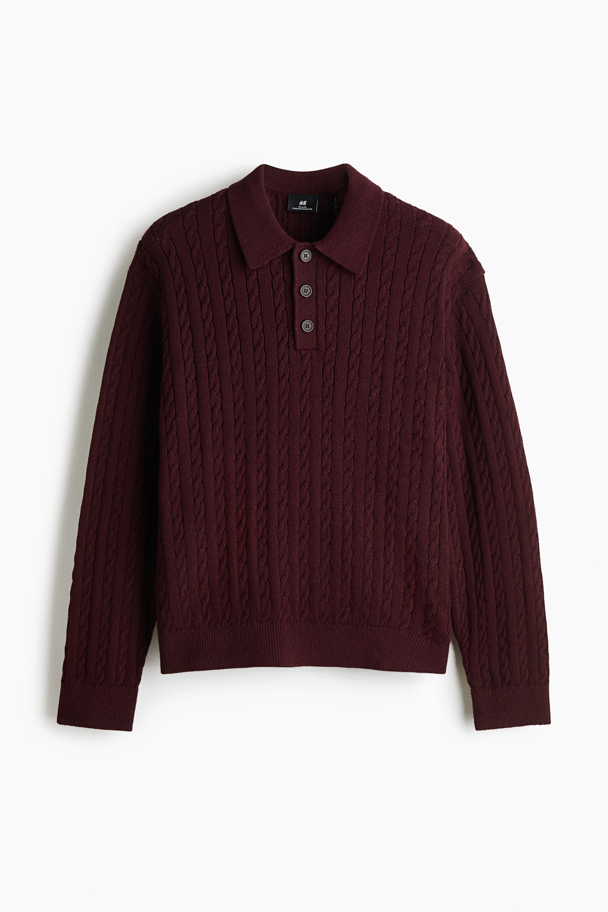 A burgundy cable-knit polo sweater is being worn by the model, featuring a ribbed collar, a button placket, dropped shoulders, and ribbed cuffs and hem. The relaxed-fit sweater is layered over a white t-shirt and paired with dark jeans, with the model keeping one hand in a pocket.