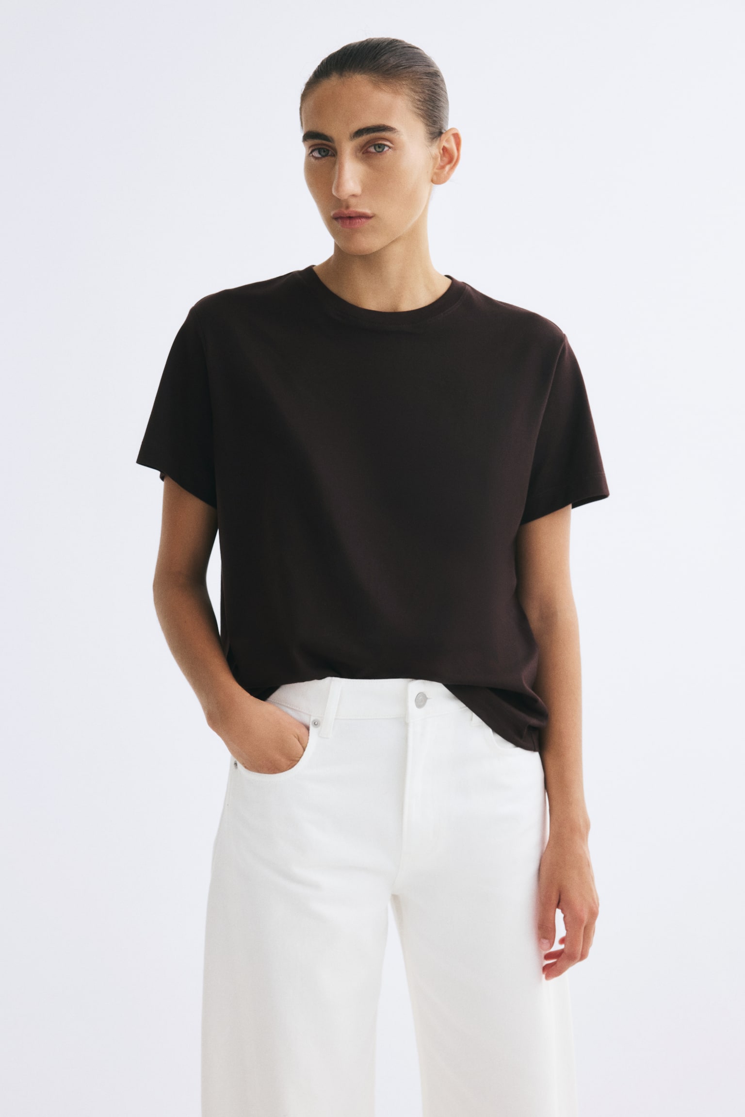 The model is wearing a dark brown short-sleeved crew neck T-shirt styled with high-waisted white jeans. She is standing with one hand resting in a pocket, showcasing the relaxed fit of the top. - 1