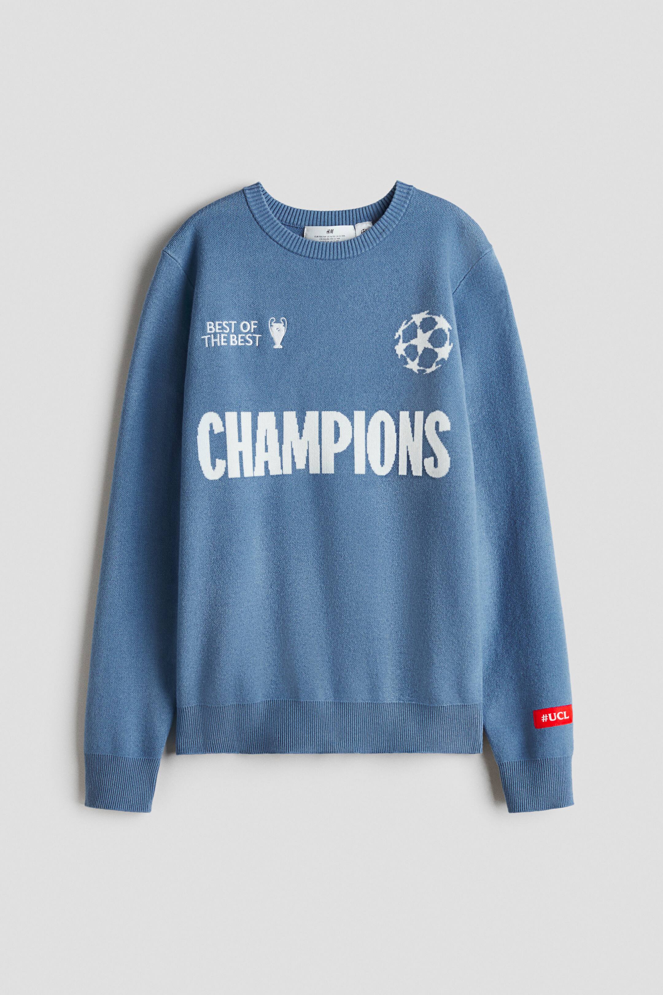 View larger image: A blue knit crewneck sweater is showcasing its front, featuring white "BEST OF THE BEST" text with a trophy icon on the upper left, a stylized football icon on the upper right, and large "CHAMPIONS" text across the chest. The sweater also highlights ribbed cuffs and hem, with a small red tag reading "#UCL" on the left sleeve cuff.