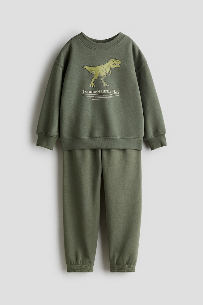 Un conjunto de dos piezas, que consiste en una sudadera verde oliva y joggers a juego, se presenta desde el FRENTE. La sudadera presenta un gráfico de Tyrannosaurus Rex en verde claro y texto en su parte delantera, un cuello redondo con ribete, hombros caídos, y puños y dobladillo acanalados. Los joggers muestran una cintura elástica y dobladillos con puños. - 1