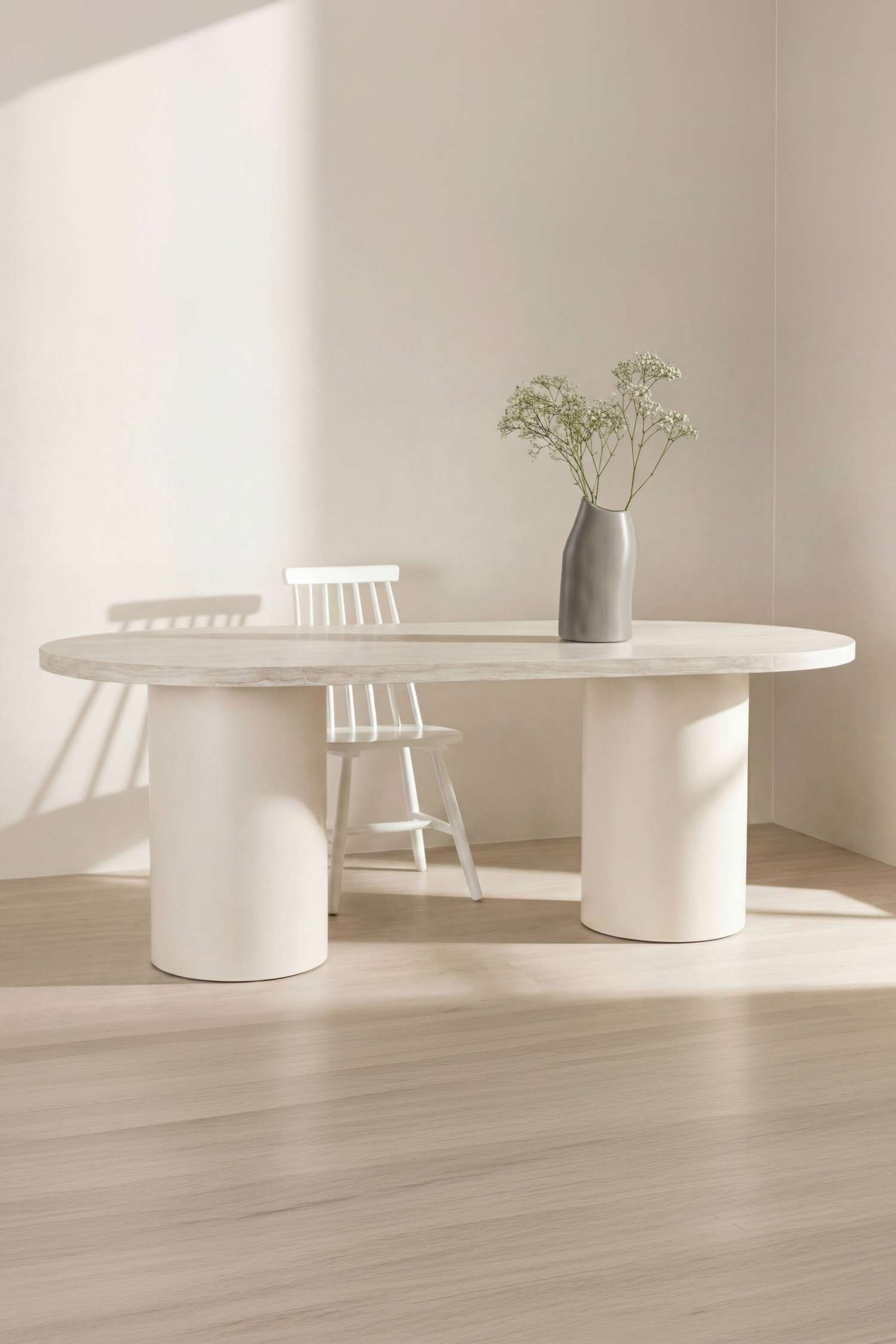 Ver imagen más grande: Una mesa de comedor ovalada presenta un tablero texturizado de color gris claro, sostenido por dos gruesas bases cilíndricas de color blanco roto. Una silla está colocada debajo, y un jarrón se encuentra sobre su superficie.