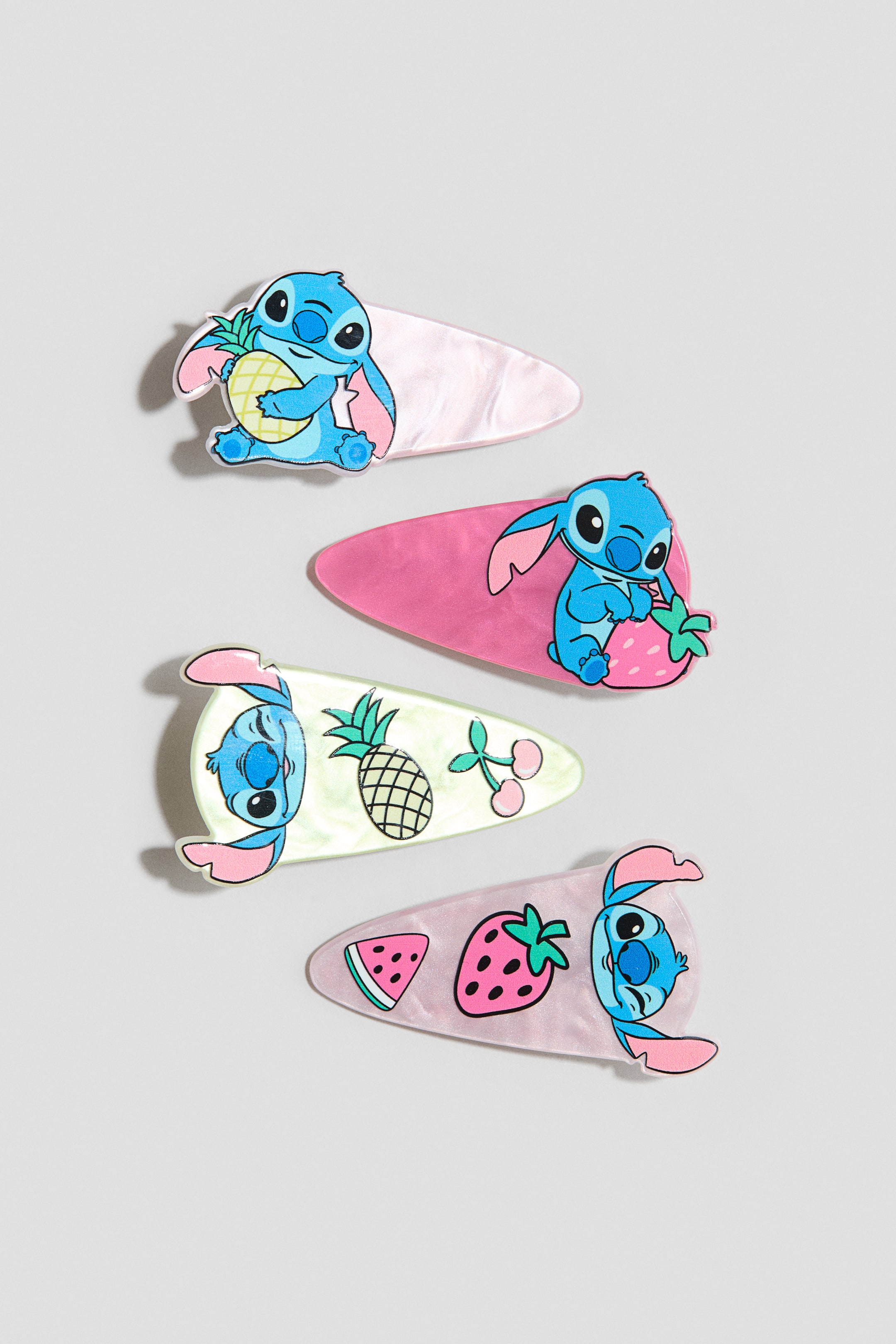 Voir une image plus grande: Paquet de 4 pinces à cheveux présentant quatre pinces de style barrette, chacune ornée d'un personnage bleu et de motifs de fruits sur le devant, avec des bases en rose pâle, rose vif, vert clair iridescent et violet clair iridescent. Le personnage bleu est montré tenant un ananas, tenant une fraise, à côté d'un ananas et de cerises, et à côté d'une tranche de pastèque et d'une fraise à travers l'ensemble.