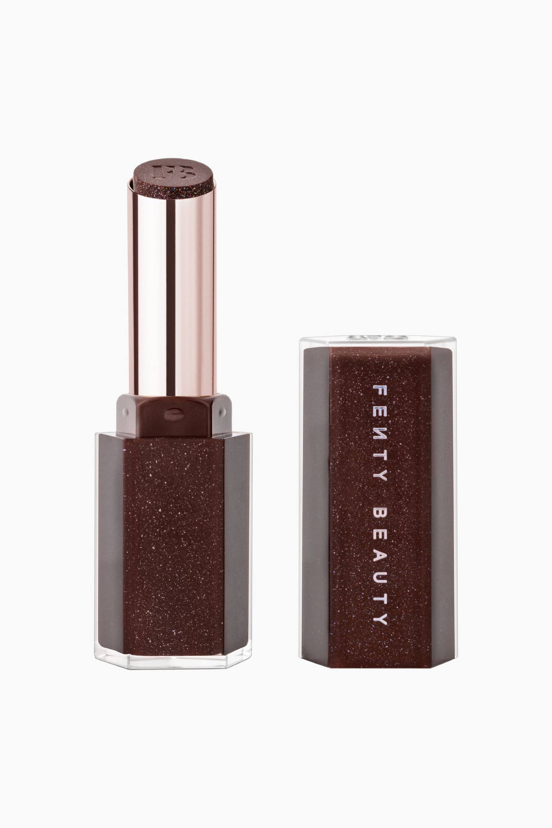 Näytä suurempi kuva: Gloss Bomb Stix High-shine Shimmer Gloss Stick seisoo pystyssä irrotetun korkkinsa vieressä, ja molemmissa osissa näkyy tumma, kimalteella höystetty kuusikulmainen muotoilu kirkkaan muovin sisällä. Avoin tikku paljastaa metallinhohtoisen ruusukultaisen sisäputken, jossa on hohtava tummanruskea gloss-syömä, johon on kohokuvioitu "FB"-logo, kun taas viereinen korkki esittää pystysuuntaista "FENTY BEAUTY" -brändäystä.