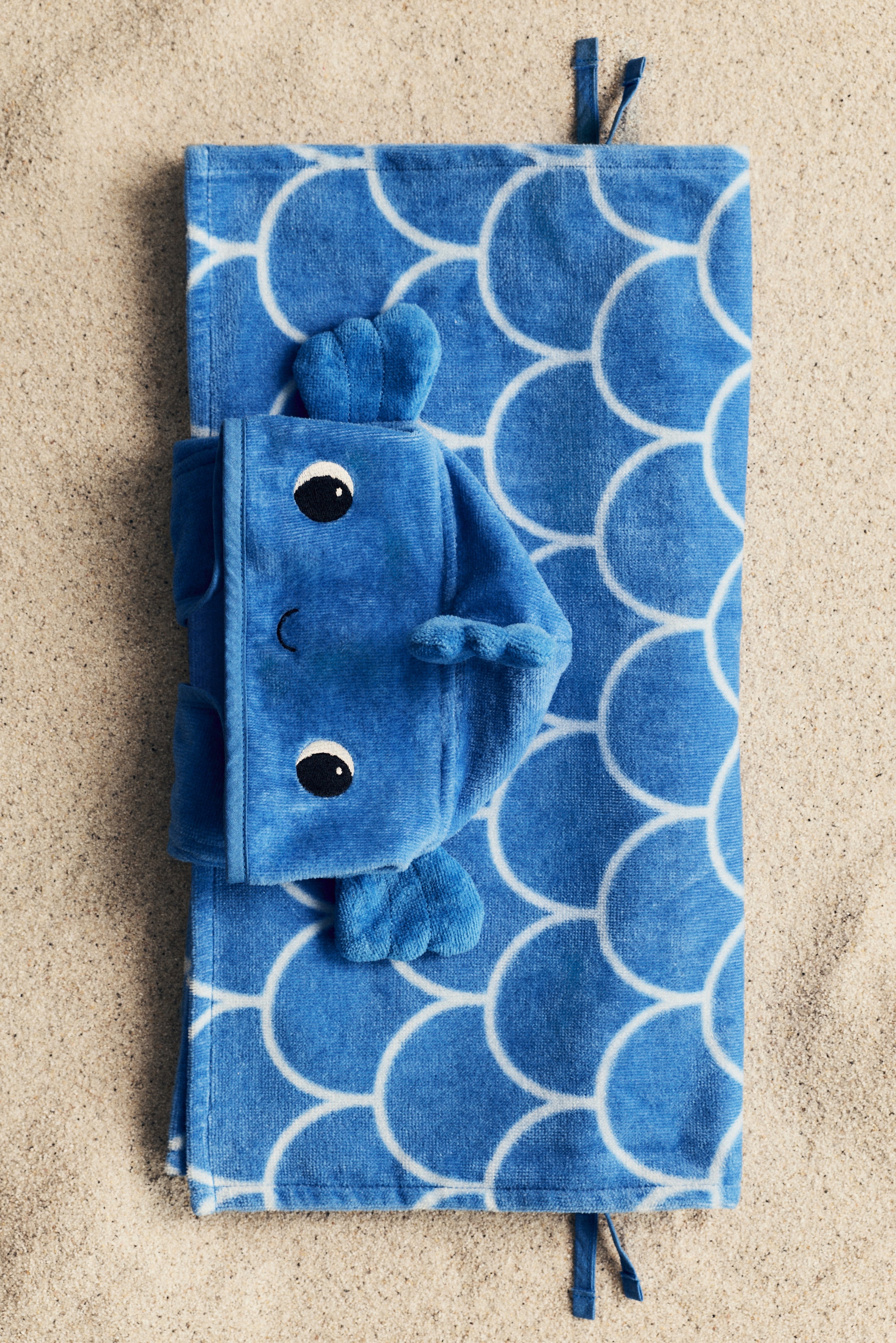 Agrandir l'image: Une cape serviette animale bleue est posée pliée, mettant en valeur son matériau en coton éponge doux avec un motif de coquilles blanches. Une capuche animale en velours bleu, ornée d'yeux noirs, d'une bouche incurvée et de petits détails semblables à des nageoires, est orientée vers l'avant sur la serviette pliée, avec des liens en tissu bleu étroit visibles aux extrémités.