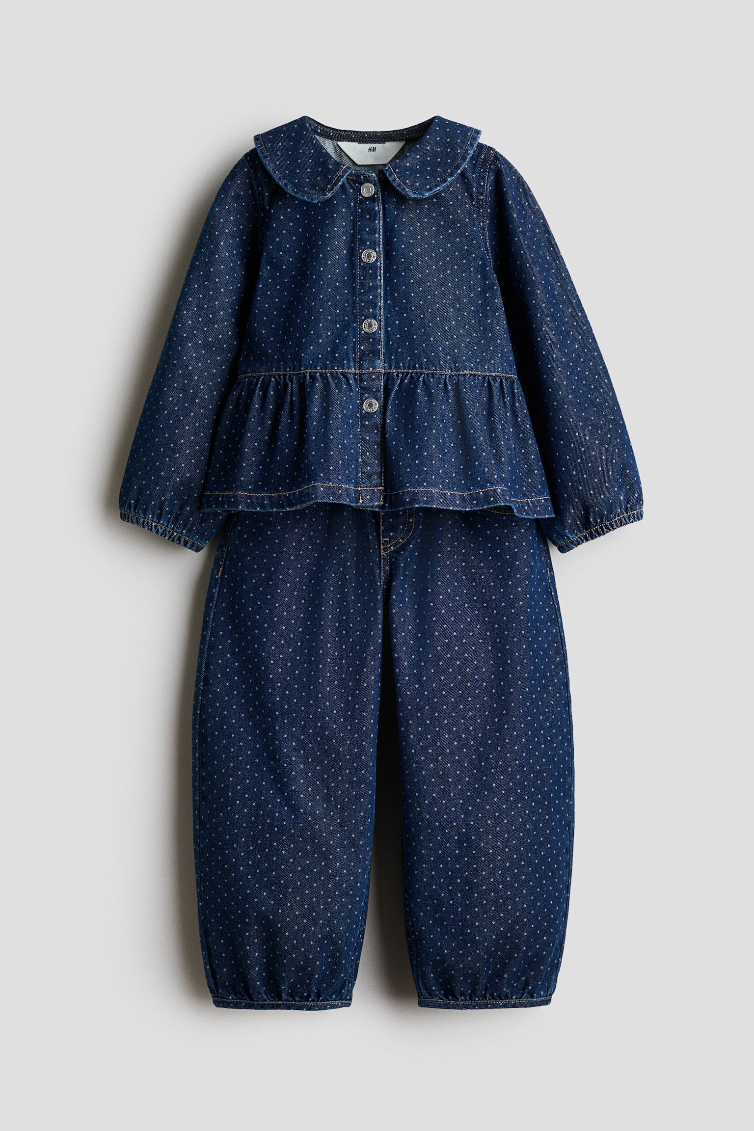 This two-piece denim set, shown from the front, features a dark blue fabric decorated with a white polka-dot pattern. The top displays a rounded Peter Pan collar, a button-up front, and a gathered peplum hem, while the matching trousers offer a relaxed fit with elasticated ankle cuffs.