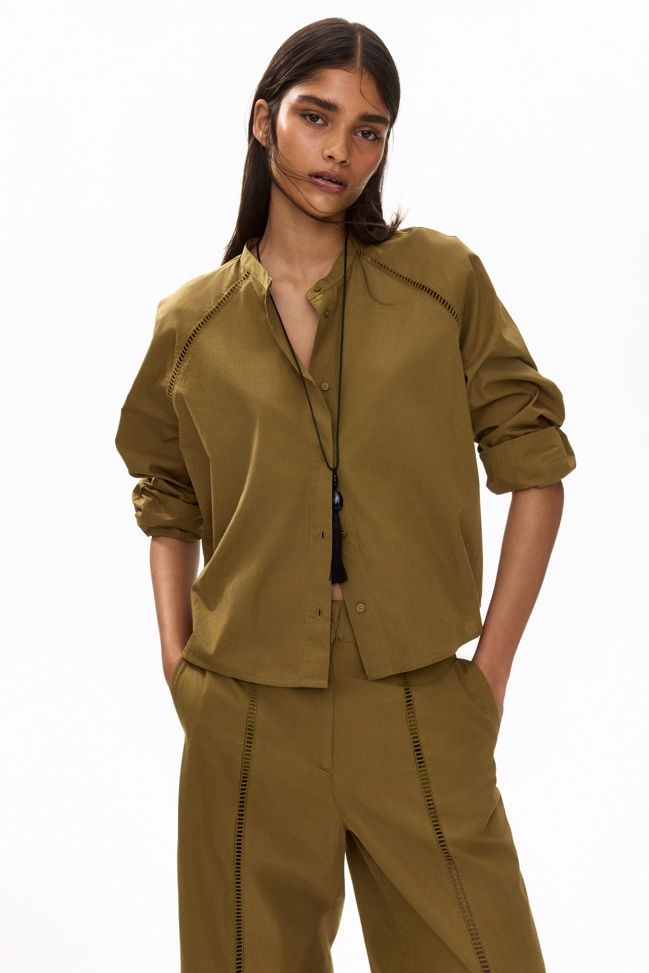 View larger image: An olive green matching shirt and trousers set is displayed front-facing. The shirt features a band collar, long sleeves with rolled-up cuffs, and cut-out detailing along the shoulder seams and collar, with its button-down front partially unbuttoned. A black long necklace with a tassel and stone pendant hangs over the shirt, and the trousers showcase vertical cut-out detailing down the outer seams.