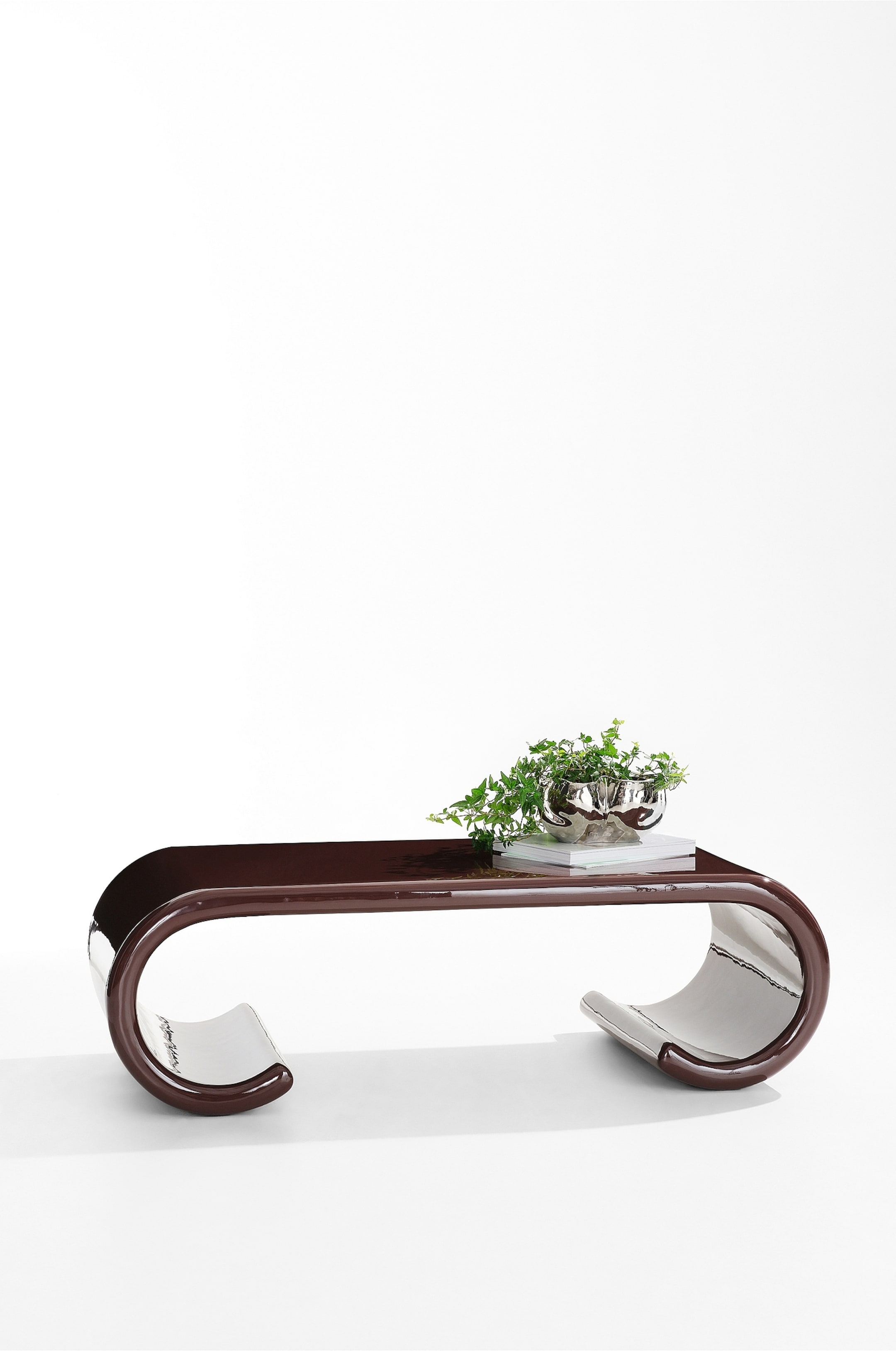 Grotere afbeelding bekijken: Een moderne salontafel, weergegeven vanuit een frontale driekwartshoek, heeft een sculpturaal ontwerp met een hoogglanzende, diepbruine afwerking en naar binnen gebogen poten in scrollvorm die een sterk reflecterende zilverkleurige onderzijde onthullen. Rechts op het vlakke tafelblad rust een kleine potplant in een metalen schaal bovenop twee boeken.
