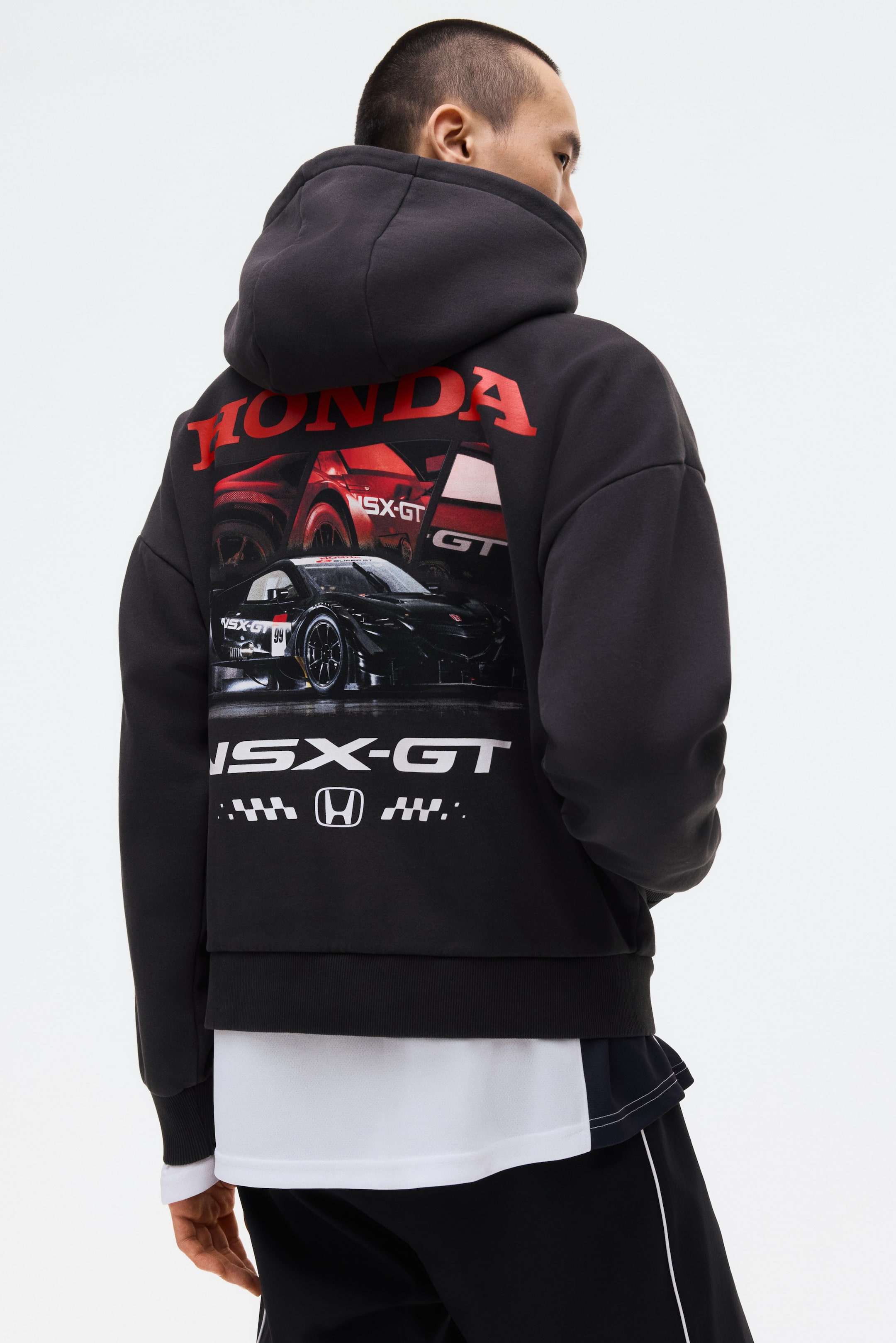 View larger image: A black hoodie is worn with its back facing the viewer, showcasing a large graphic print. The print features the red 'HONDA' logo, a red and black NSX-GT race car design, and 'NSX-GT' text alongside a Honda logo and checkered pattern at the bottom, with a white t-shirt layered beneath the hoodie's ribbed hem.