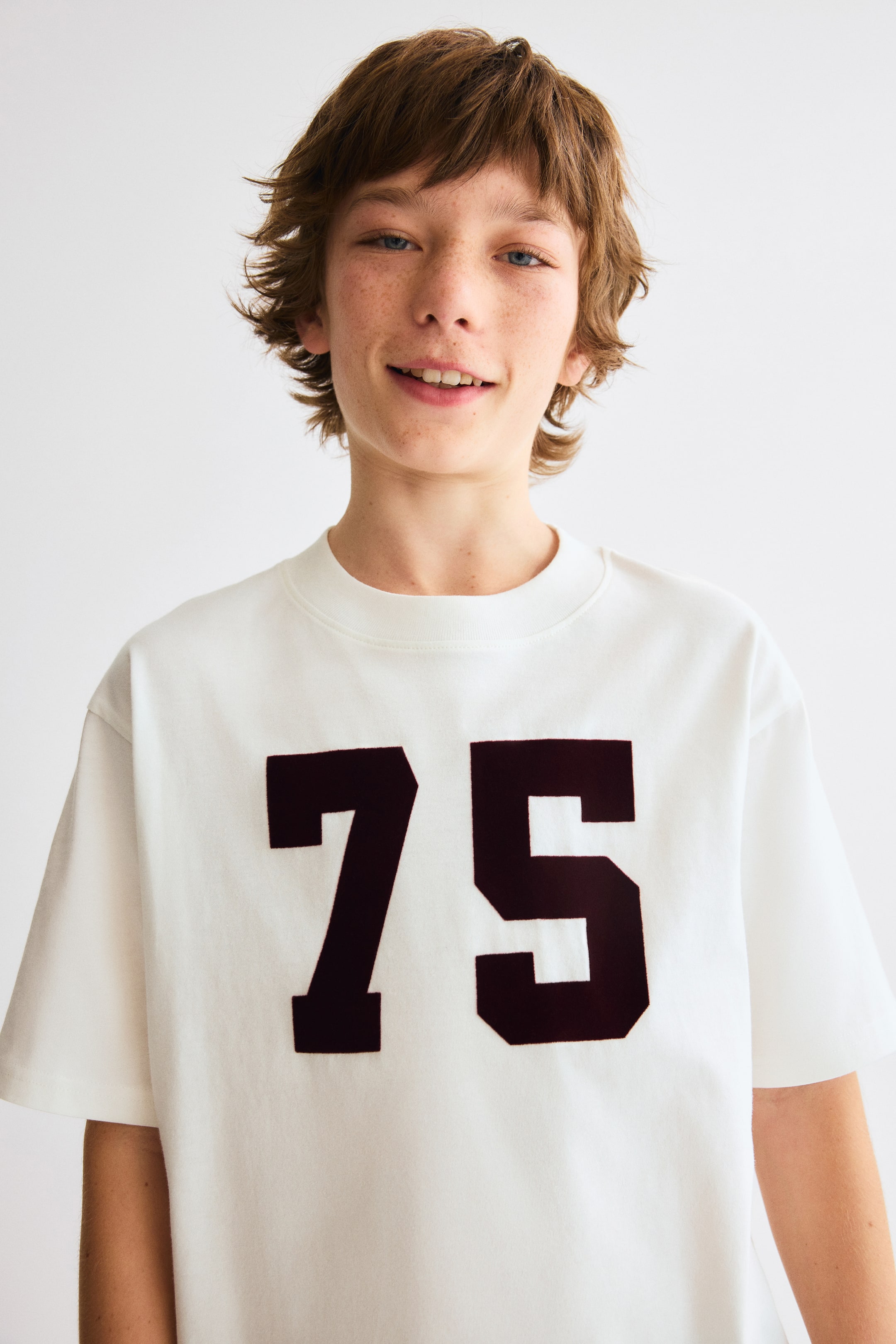 Prikaži veću sliku: Bela majica sa okruglim izrezom ima velike, tamno bordo brojeve '75' istaknute na prednjoj strani, kratke rukave i opušten kroj.