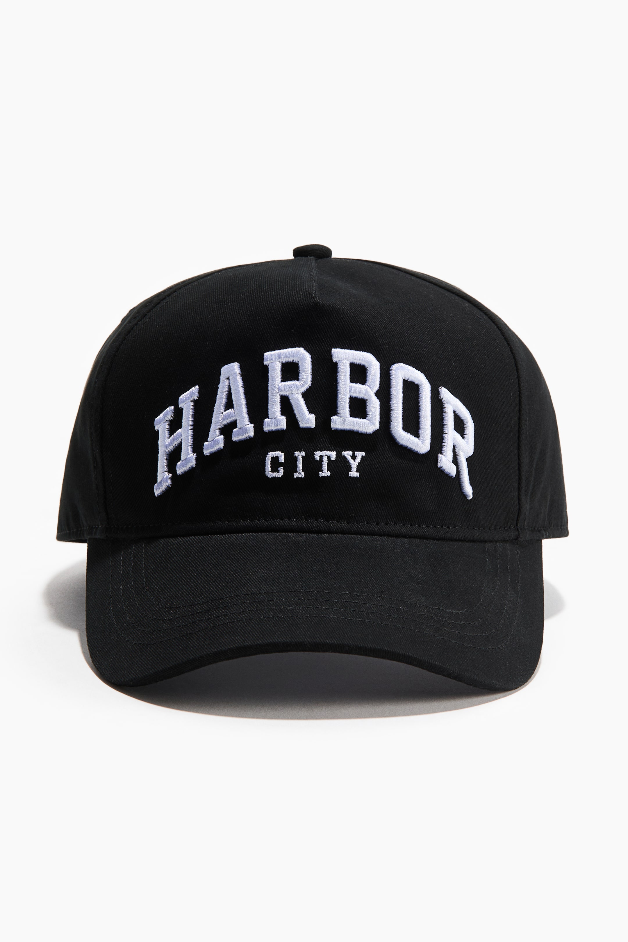 Zobraziť väčší obrázok: Čierna baseballová čiapka je otočená dopredu, zobrazujúca biely vyšívaný text „HARBOR“ v oblúku s „CITY“ umiestneným pod ním. Čiapka má zakrivený šilt s viditeľným prešívaním a malým čiernym gombíkom na vrchole.