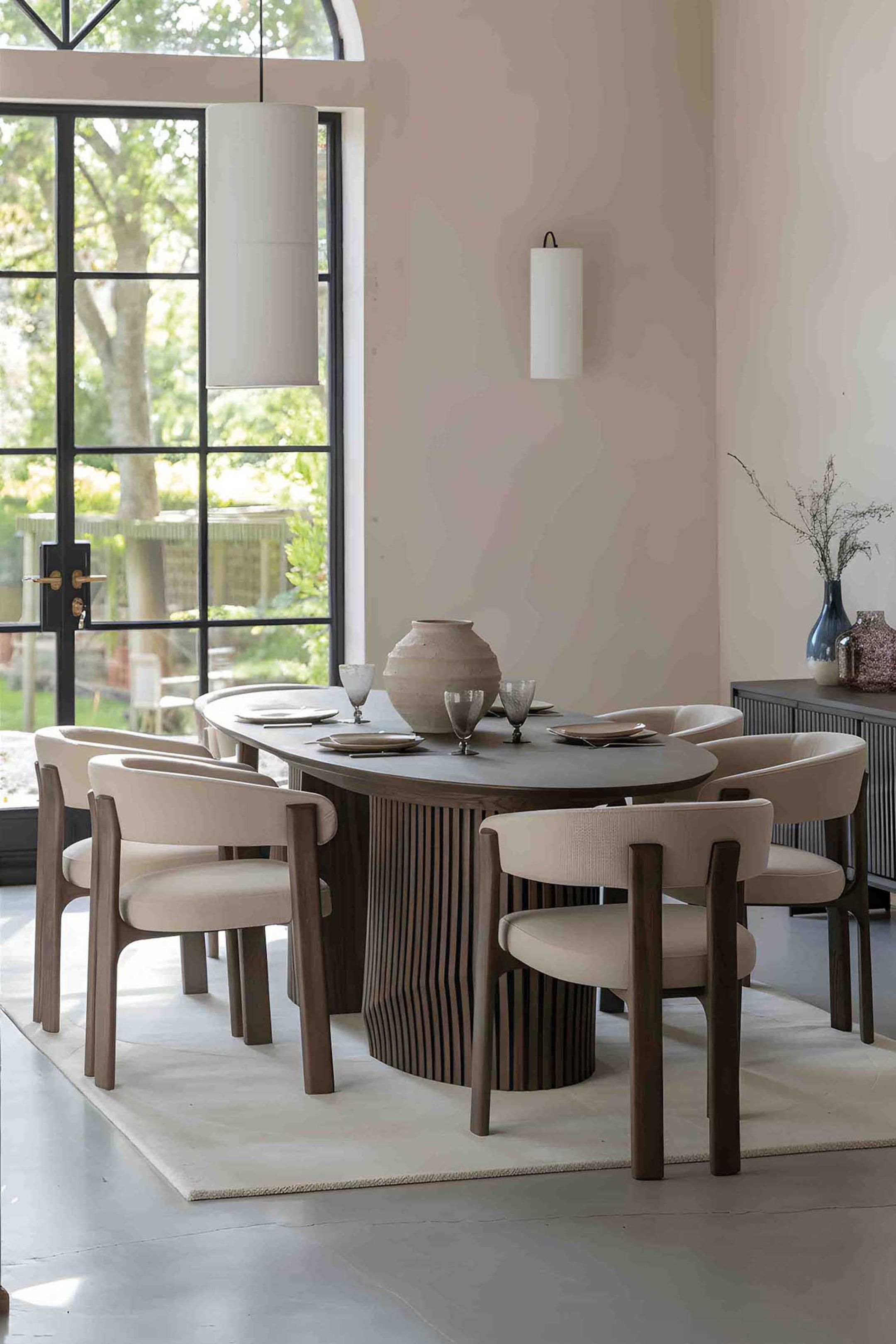 View larger image: The dining table features an elongated oval top with a dark finish, resting on a central pedestal base with a vertical slatted design, and is set with plates, glasses, and a large textured vase.