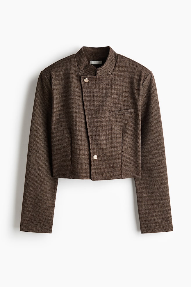 Eine kurz geschnittene, strukturierte braune Jacke mit feinem Karomuster und asymmetrischem Frontverschluss mit zwei runden MetallknÃ¶pfen. Die Jacke verfÃ¼gt Ã¼ber einen hohen Stehkragen, ausgeprÃ¤gte Schultern, eine einzelne Brust-Leistentasche und lange, gerade Ãrmel. - 1