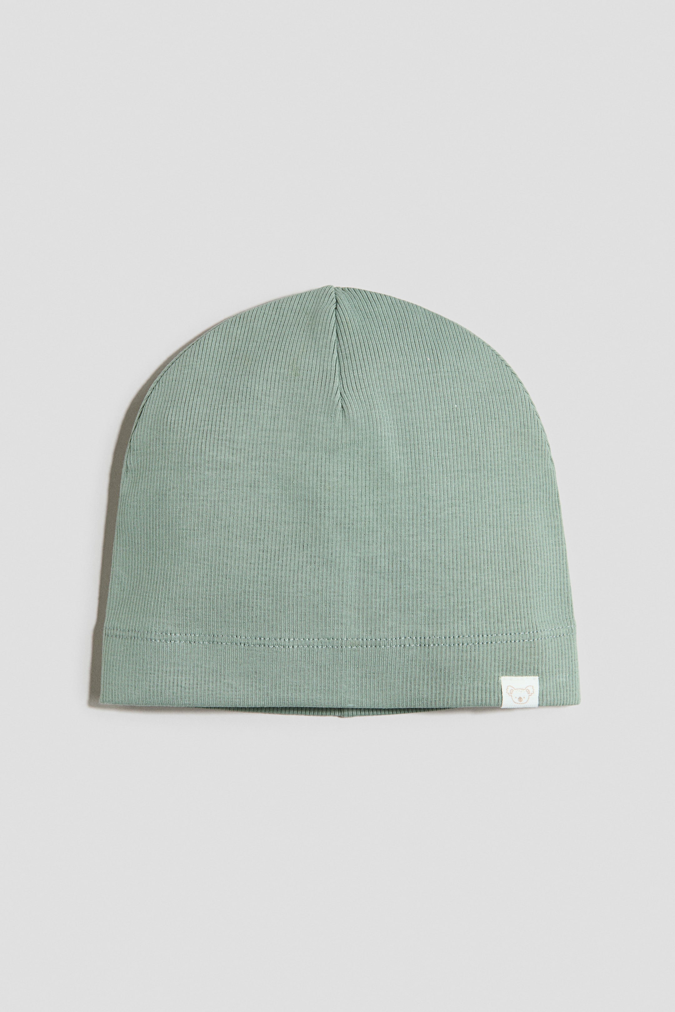 Ver imagen más grande: Un gorro verde claro está colocado plano y orientado hacia el frente, con textura acanalada de punto jersey en toda la prenda. Una pequeña etiqueta tejida blanca con un emblema de tono claro está fijada en la parte frontal, en el lado inferior derecho del borde.