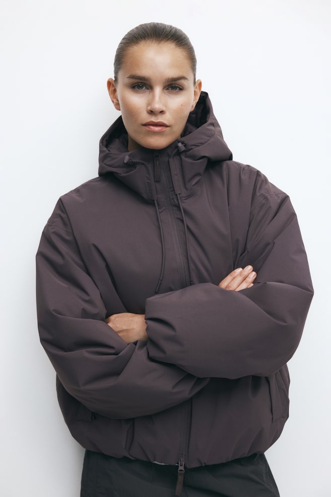 A dark plum-colored hooded puffer jacket is being worn by the model, who is posing with crossed arms. The jacket features a high-neck hood with adjustable drawstrings, a front zipper, and voluminous sleeves. - 1