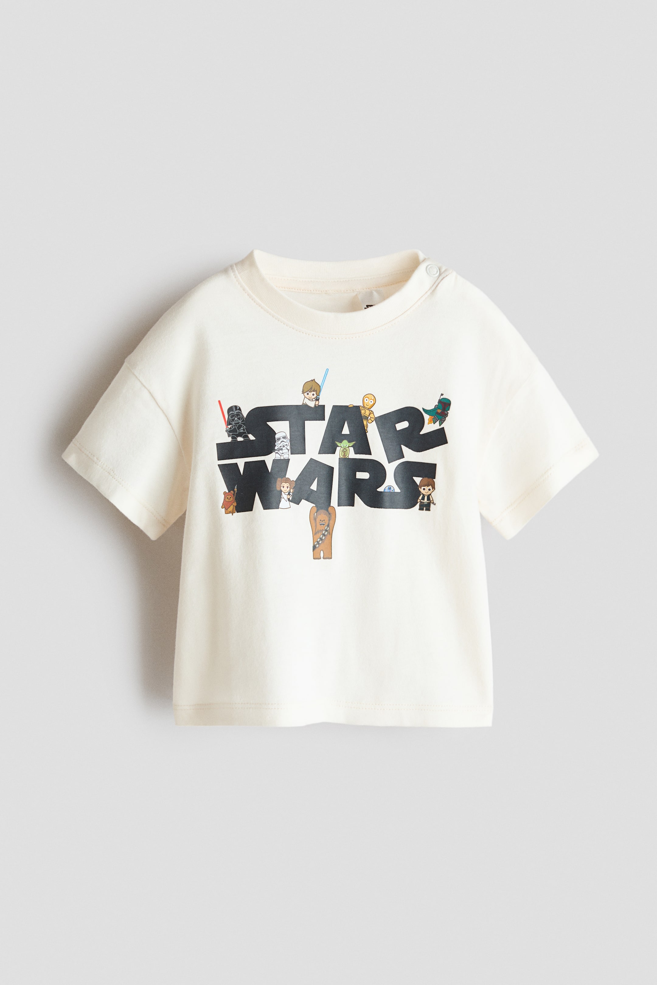 View larger image: The printed cotton T-shirt comes in a cream hue, showcasing a ribbed crew neck and short sleeves. Two snap buttons secure the right shoulder, while a prominent dark grey 'STAR WARS' logo adorns the front, encircled by small, stylized characters.