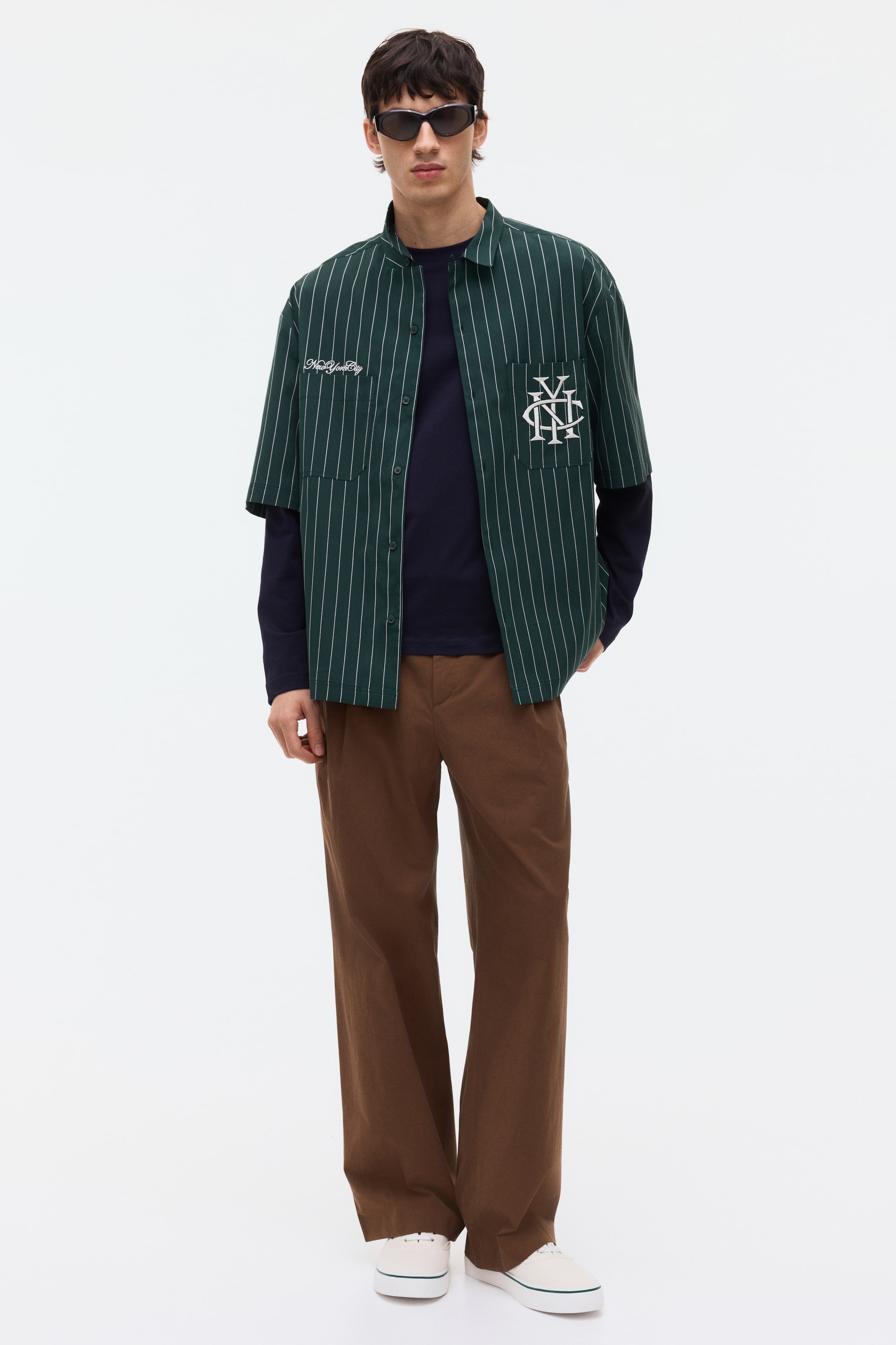 Ver imagen más grande: El producto es un conjunto para hombre, de frente, que presenta una camisa de manga corta verde oscuro con rayas verticales blancas, el texto "New York City" y un monograma bordado. Esta camisa se lleva sobre una camiseta de manga larga azul marino lisa, y el conjunto se completa con unos pantalones de pierna ancha marrón oscuro y zapatillas de lona blancas con una franja verde en la suela.