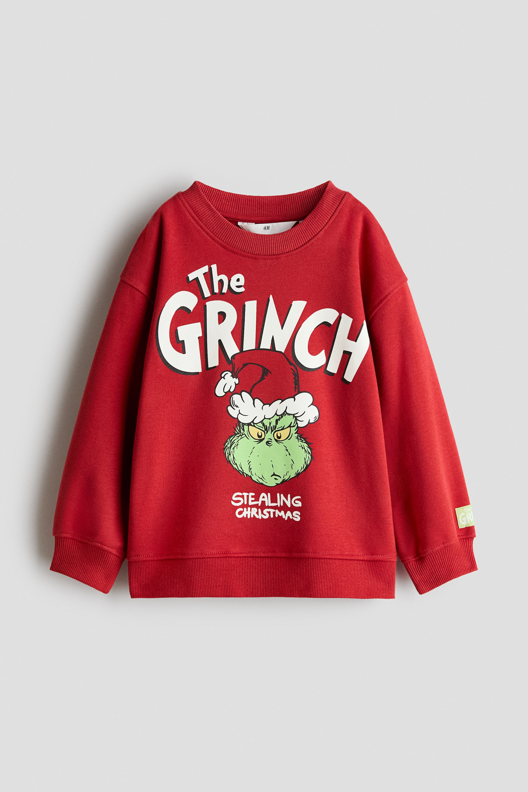 View larger image: A red sweatshirt is shown from the front, featuring a graphic print of The Grinch wearing a Santa hat, with the text "The GRINCH" and "STEALING CHRISTMAS" across the chest. The design includes a round ribbed neckline, dropped shoulders, long sleeves, and ribbed cuffs and hem.