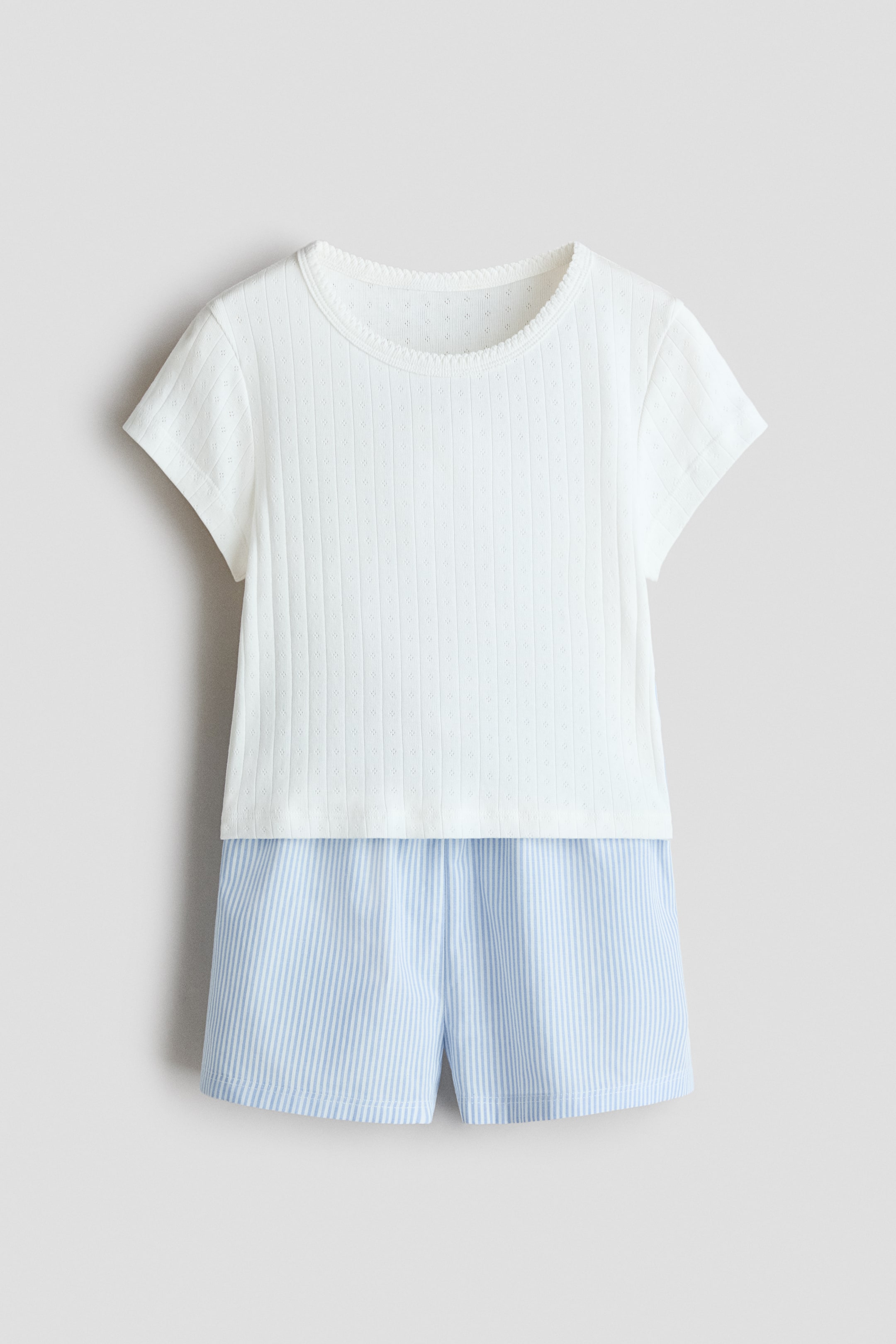 Grotere afbeelding bekijken: Een pyjamaset wordt van voren gepresenteerd, met een witte top met korte mouwen en een geribbelde textuur, geperforeerde details en een geschulpte ronde hals, gecombineerd met blauw-wit verticaal gestreepte shorts.