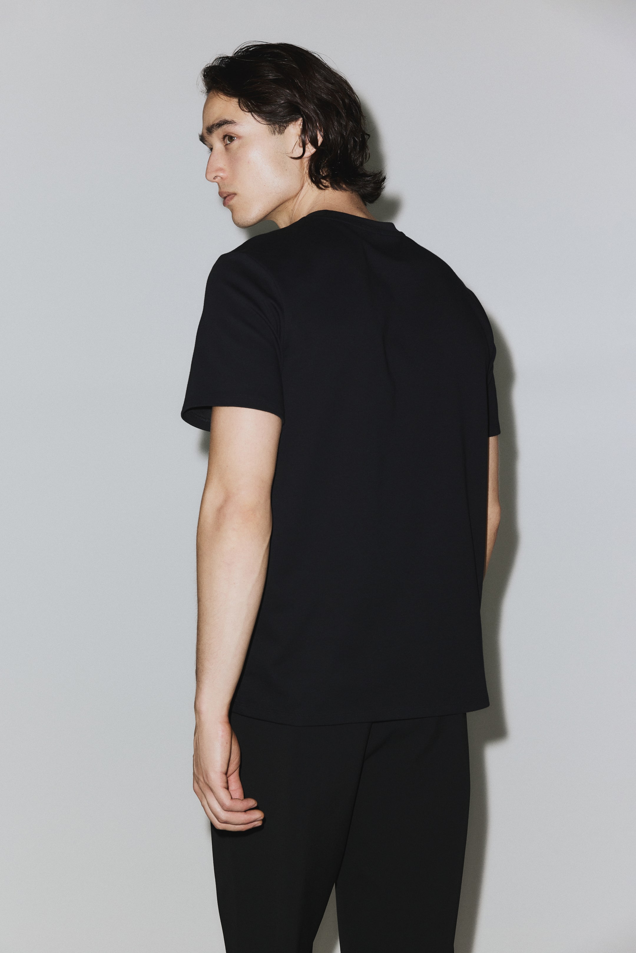 Größeres Bild ansehen: Ein schwarzes, kurzärmeliges T-Shirt wird aus einer schräg hinten-rechten Perspektive präsentiert. Es zeigt einen Rundhalsausschnitt, liegt glatt über Rücken und Schultern und endet in einem geraden Saum.