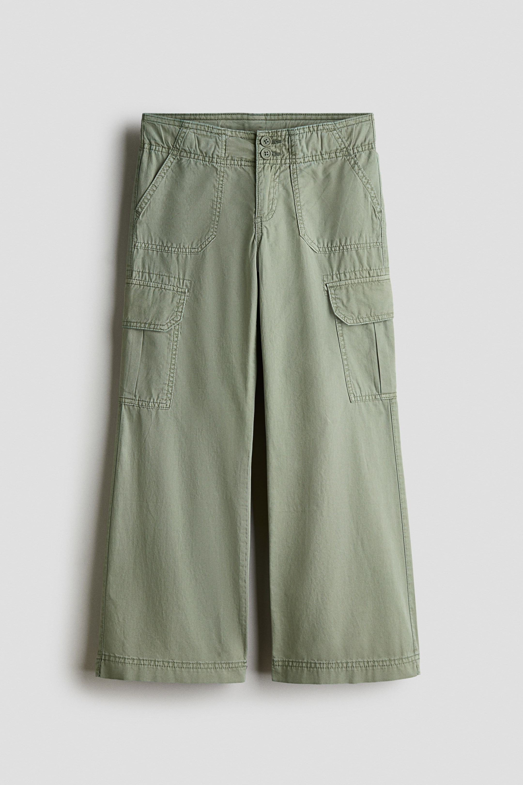 Grotere afbeelding bekijken: Een paar saliegroene cargobroeken hangt met de voorkant naar voren en toont een ontspannen wide-leg silhouet en een dubbele knoopsluiting aan de tailleband. De broek is voorzien van gebogen opgestikte zakken aan de voorkant, cargo-zakken aan de zijkant met kleppen en opvallende sierstiksels langs de naden.