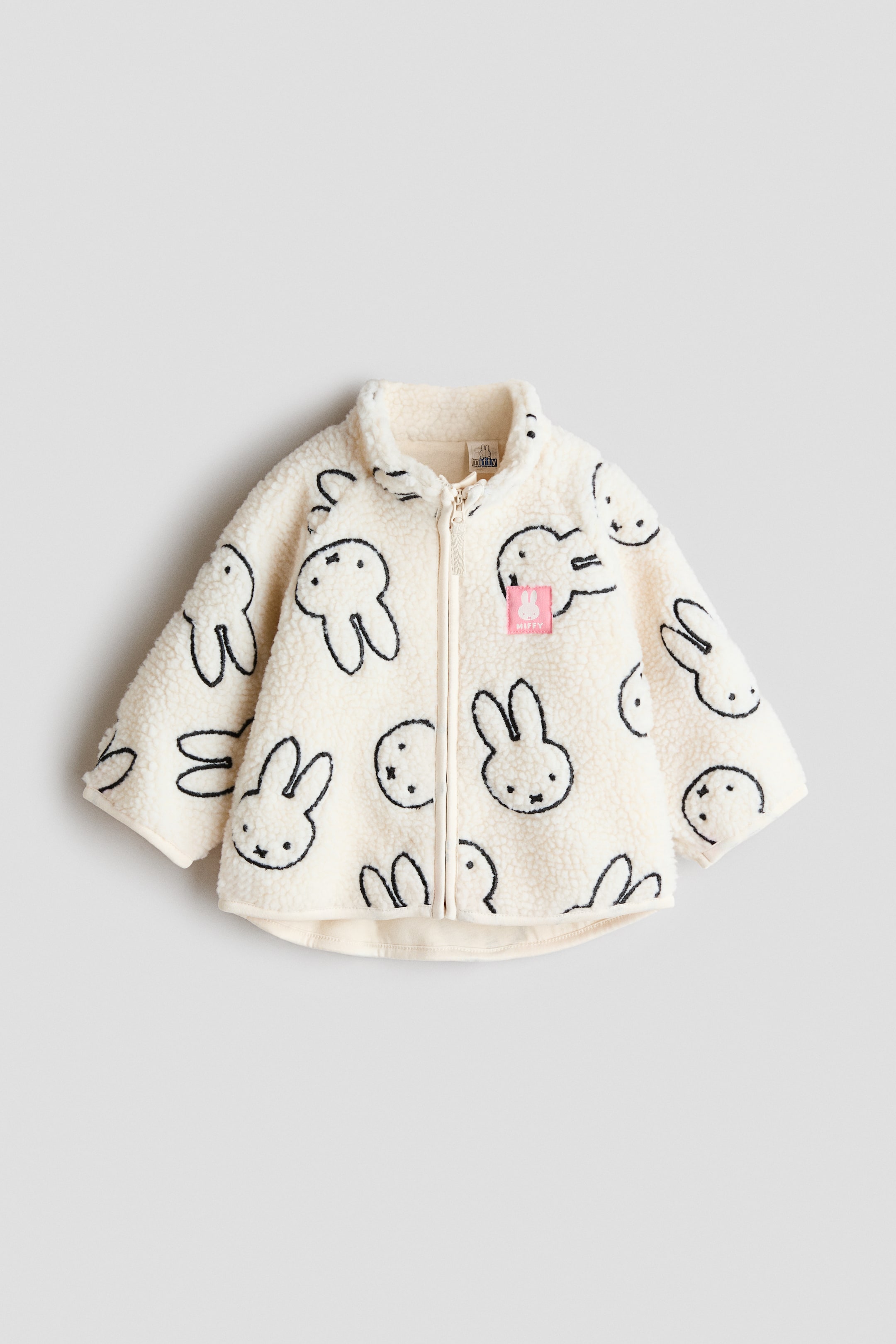 View larger image: A cream-coloured sherpa jacket featuring a full front zip, a standing collar, and an all-over pattern of black Miffy bunny outlines, with a small pink Miffy label positioned on the right chest.