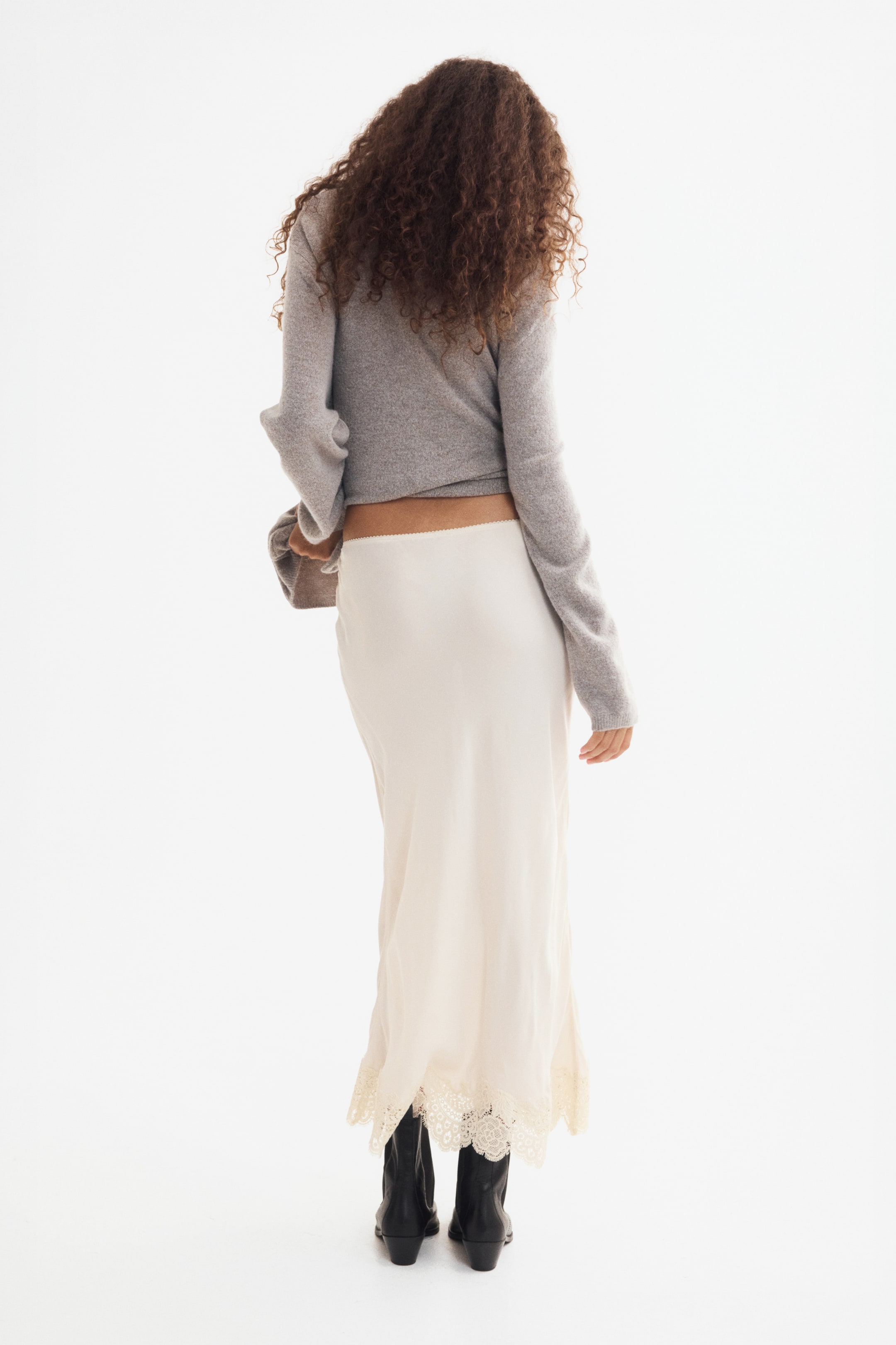 View larger image: A cream-colored, calf-length skirt in an airy viscose weave is worn by the model, shown from behind. It features an elasticated waist, visible above the hem of a gray sweater, and a scalloped lace trim at the hem, flowing over black boots.