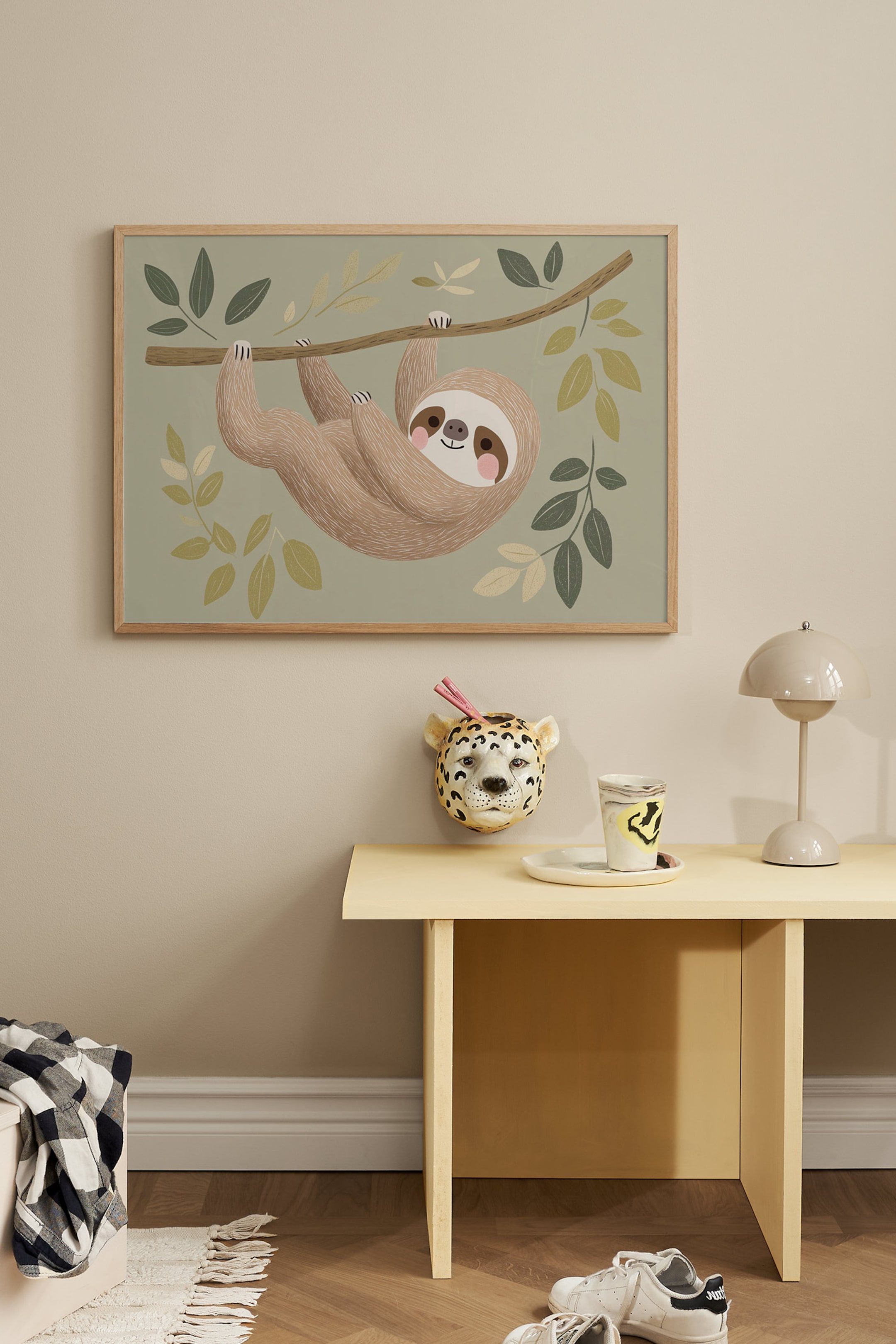 Ver imagem maior: Uma impressão artística emoldurada retangular apresenta uma ilustração de desenho animado de uma preguiça castanha sorridente pendurada num ramo de árvore. A obra de arte destaca um fundo verde-sálvia decorado com folhas estilizadas em tons de verde-escuro, oliva e bege, tudo enquadrado por uma moldura fina de madeira de tom claro.