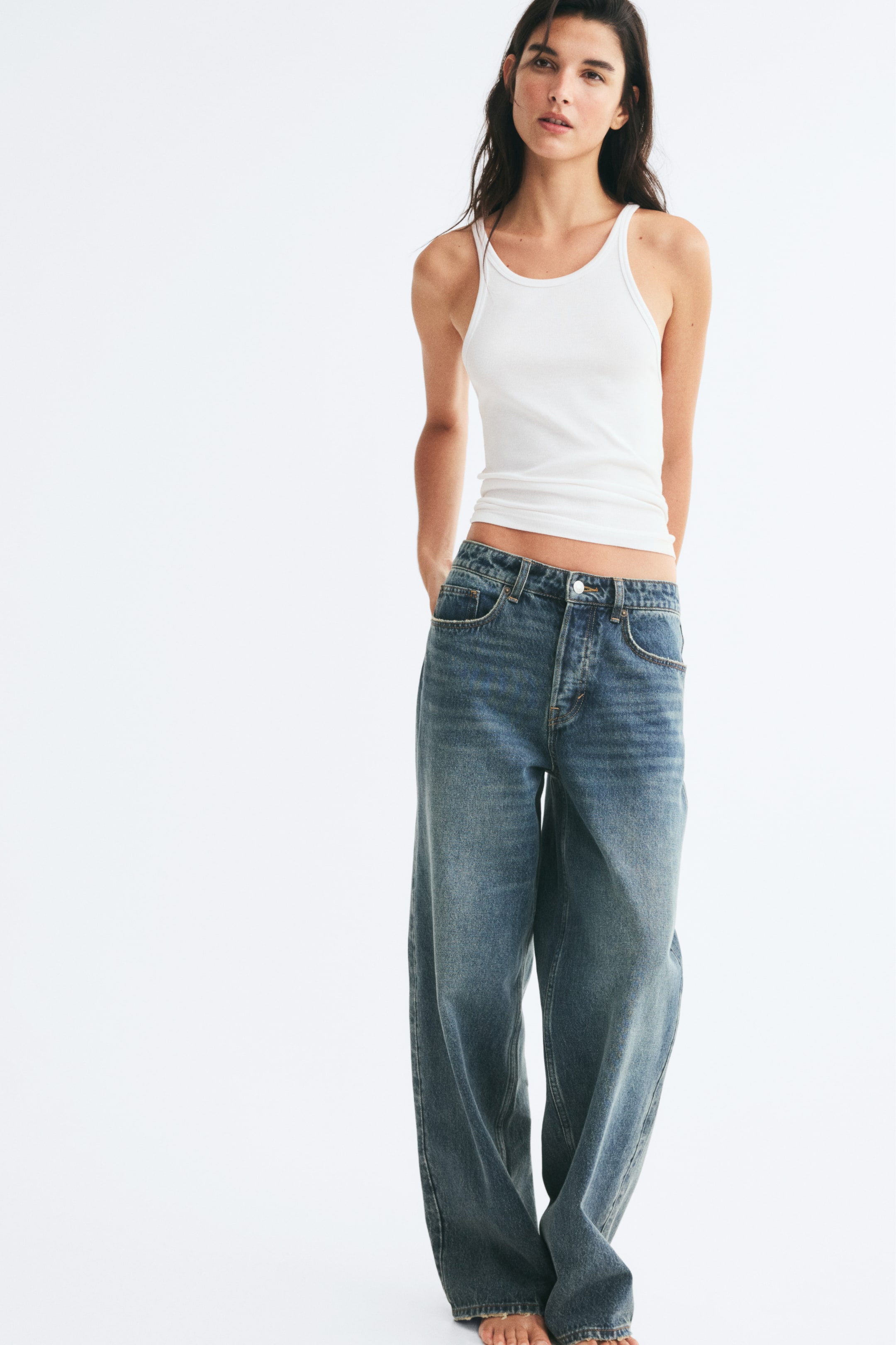 View larger image: The model is wearing blue denim jeans with a regular waist and a loose, baggy fit, standing with one hand resting in a front pocket. The five-pocket jeans feature a faded medium wash and are paired with a white ribbed tank top.