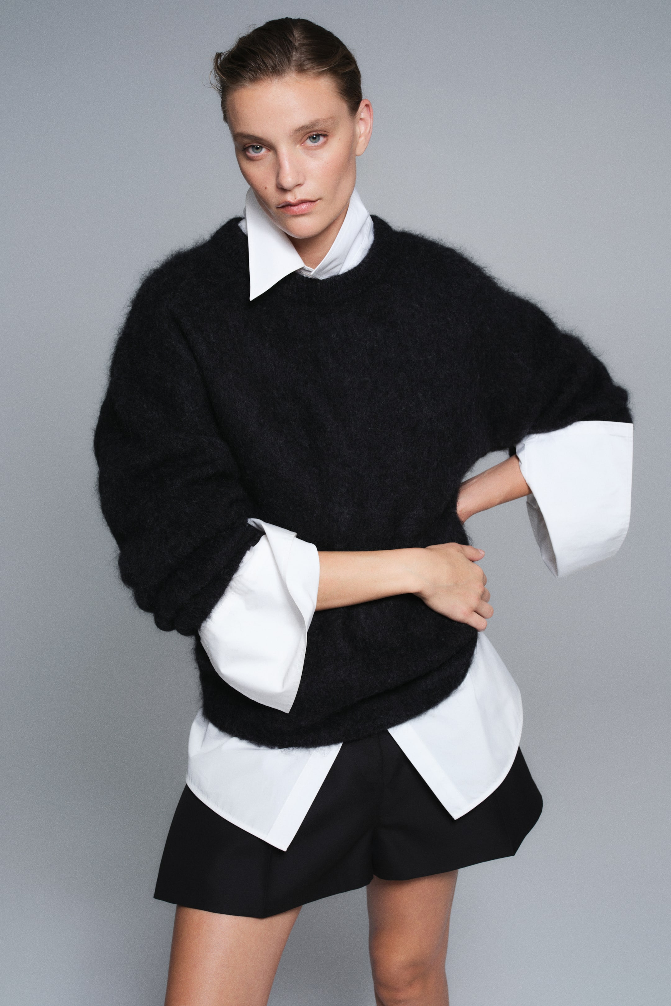 View larger image: An oversized black sweater in a fuzzy, textured knit is shown from the front, featuring dropped shoulders and a ribbed crew neckline. The sweater is layered over a structured white collared shirt, with prominent cuffs and hem peeking out to emphasise its relaxed, slouchy fit.