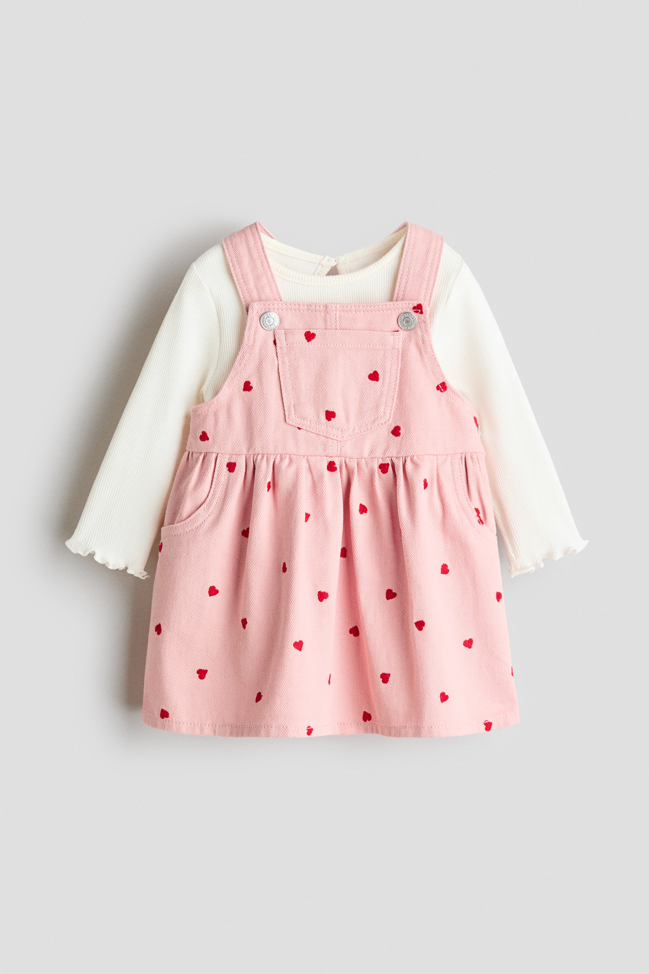 View larger image: A pink overall dress, adorned with small red hearts, is layered over a cream-colored, long-sleeved ribbed shirt. The dress features adjustable shoulder straps with silver-toned button closures, a bib pocket, and a gathered skirt with side pockets; the shirt highlights ruffled cuffs.