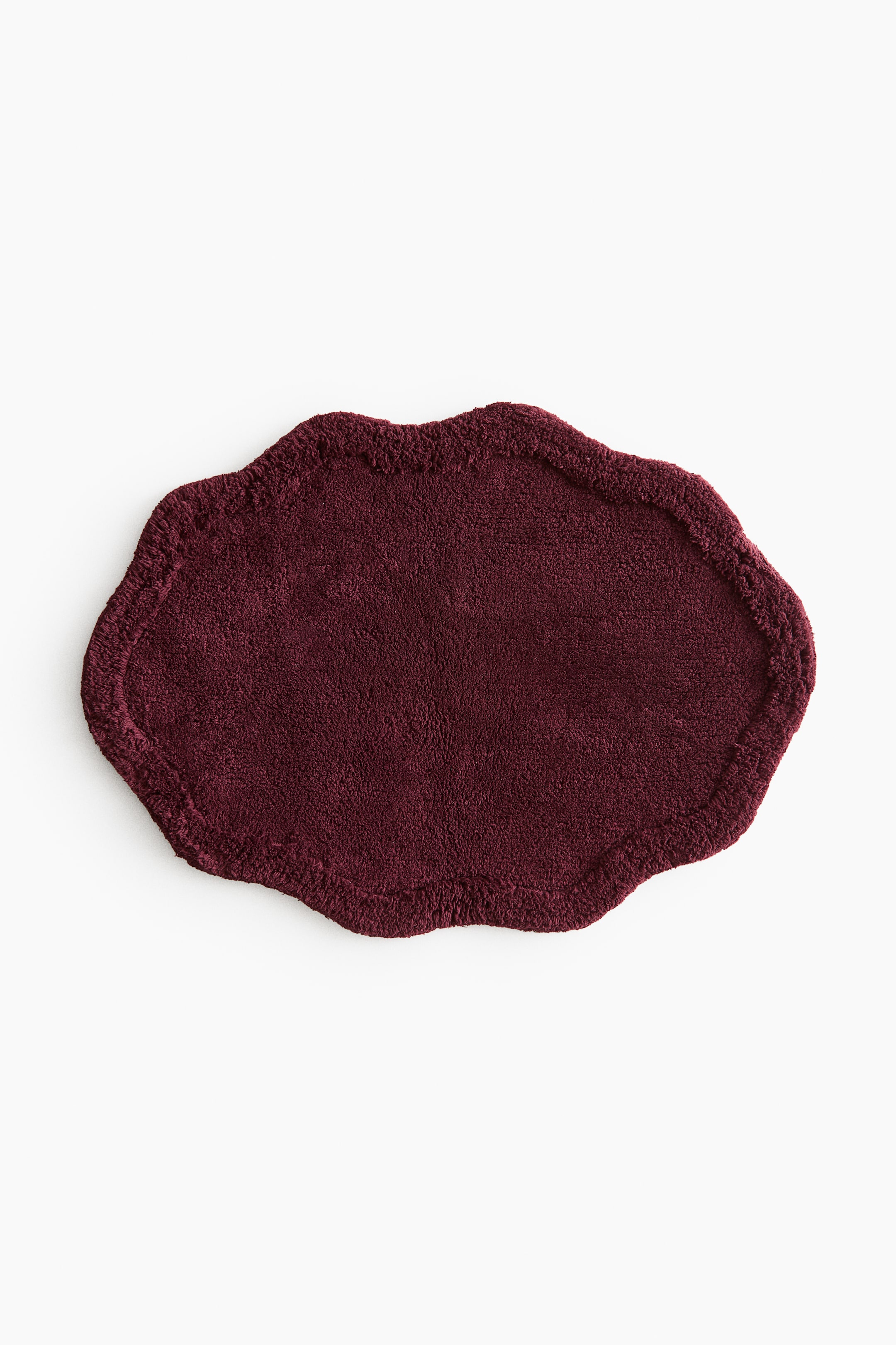 View larger image: A dark red tufted cotton bath mat is lying flat, displaying an irregular, wavy silhouette and a distinct raised border framing its edges.