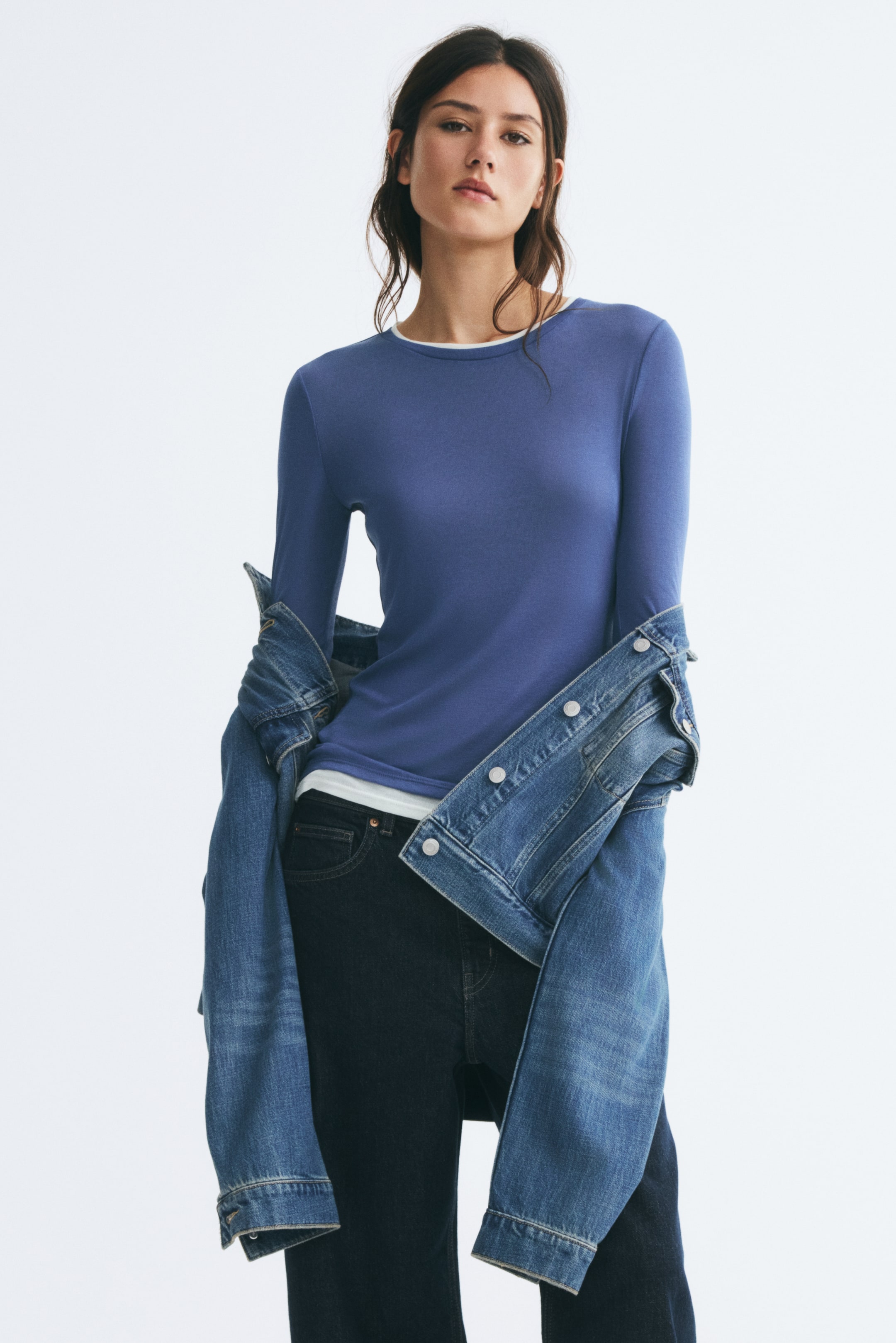 View larger image: A blue long-sleeve knit top with white accent trims at the crew neckline and hem is worn by the model, creating a layered look. The top is styled with dark denim jeans and a blue denim jacket draped off the shoulders.