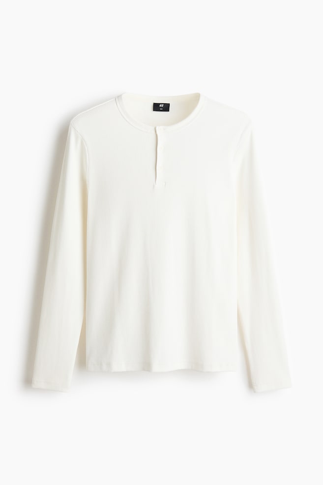 Una camiseta de punto acanalado de manga larga en color blanco roto, orientada hacia adelante, con cuello redondo y una tapeta Henley de tres botones. - 1