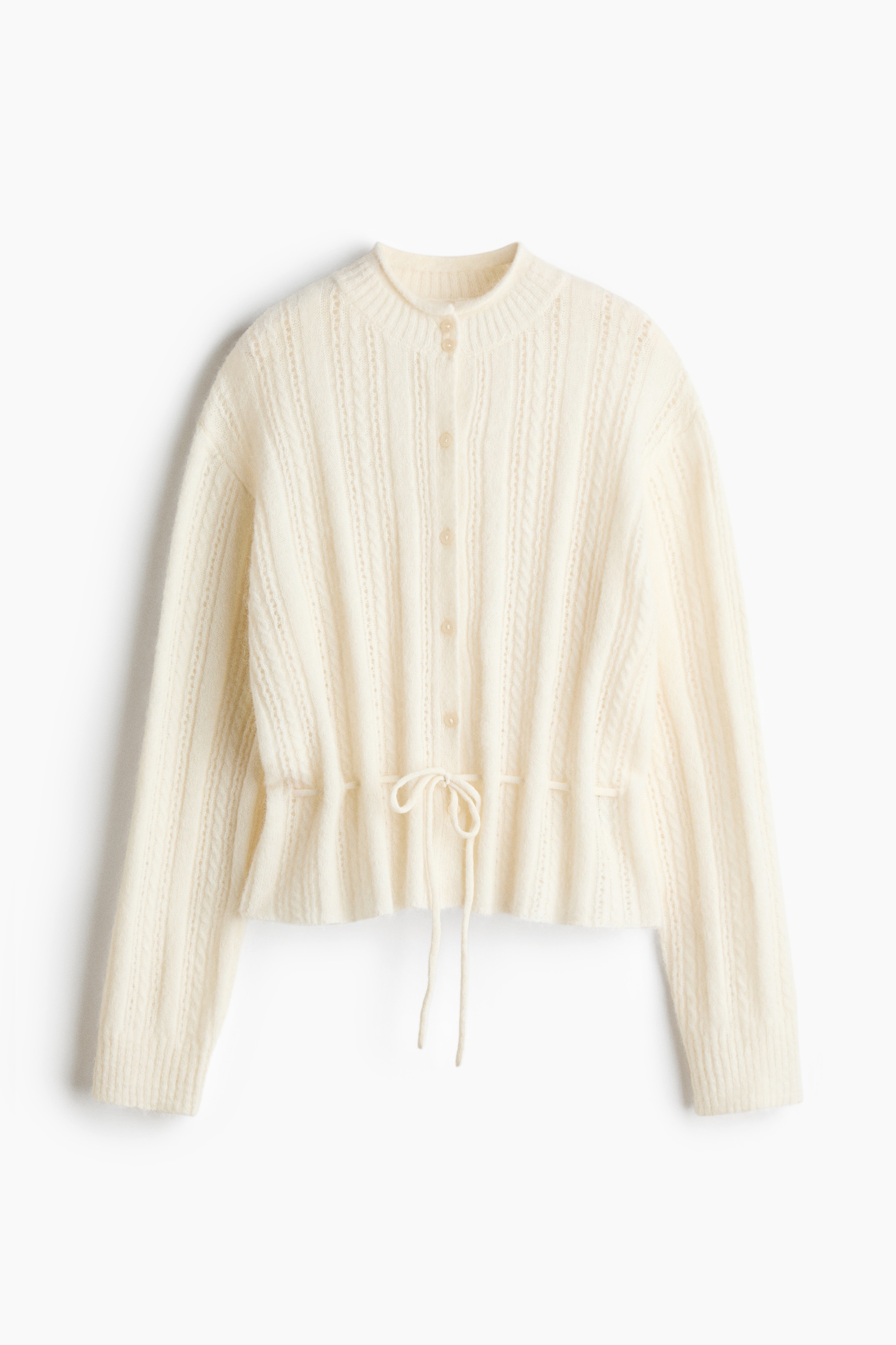 An off-white pointelle and cable-knit cardigan is being worn by the model, featuring a high ribbed neckline, long sleeves, and a drawstring tied at the waist. The cardigan is paired with dark brown leather-like trousers.