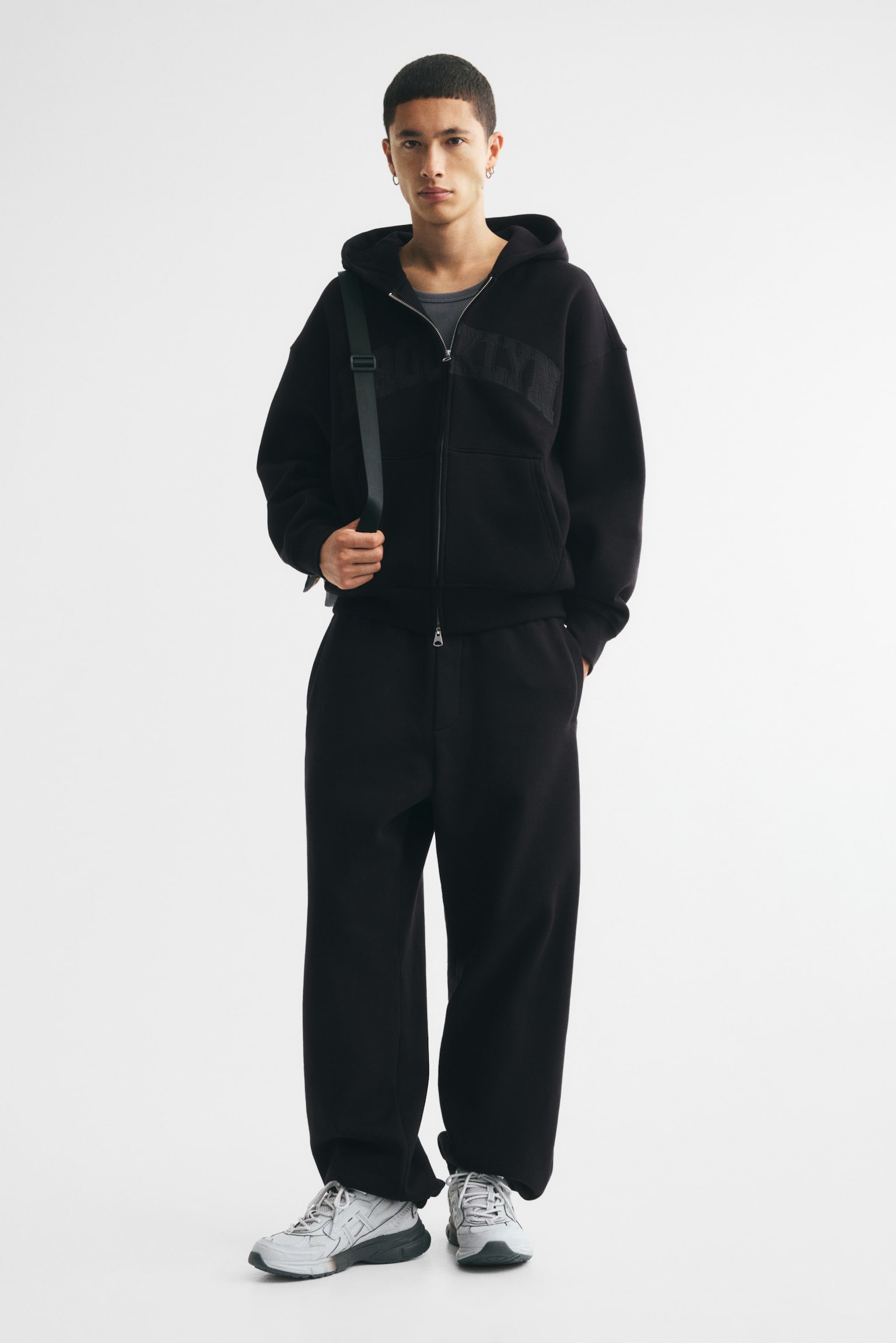 A black full-zip hoodie is facing front, featuring a silver-tone zip, a hood, and "BROOKLYN" appliquéd in matching black across the chest. It showcases ribbed cuffs, a ribbed hem, and two front patch pockets. - 1
