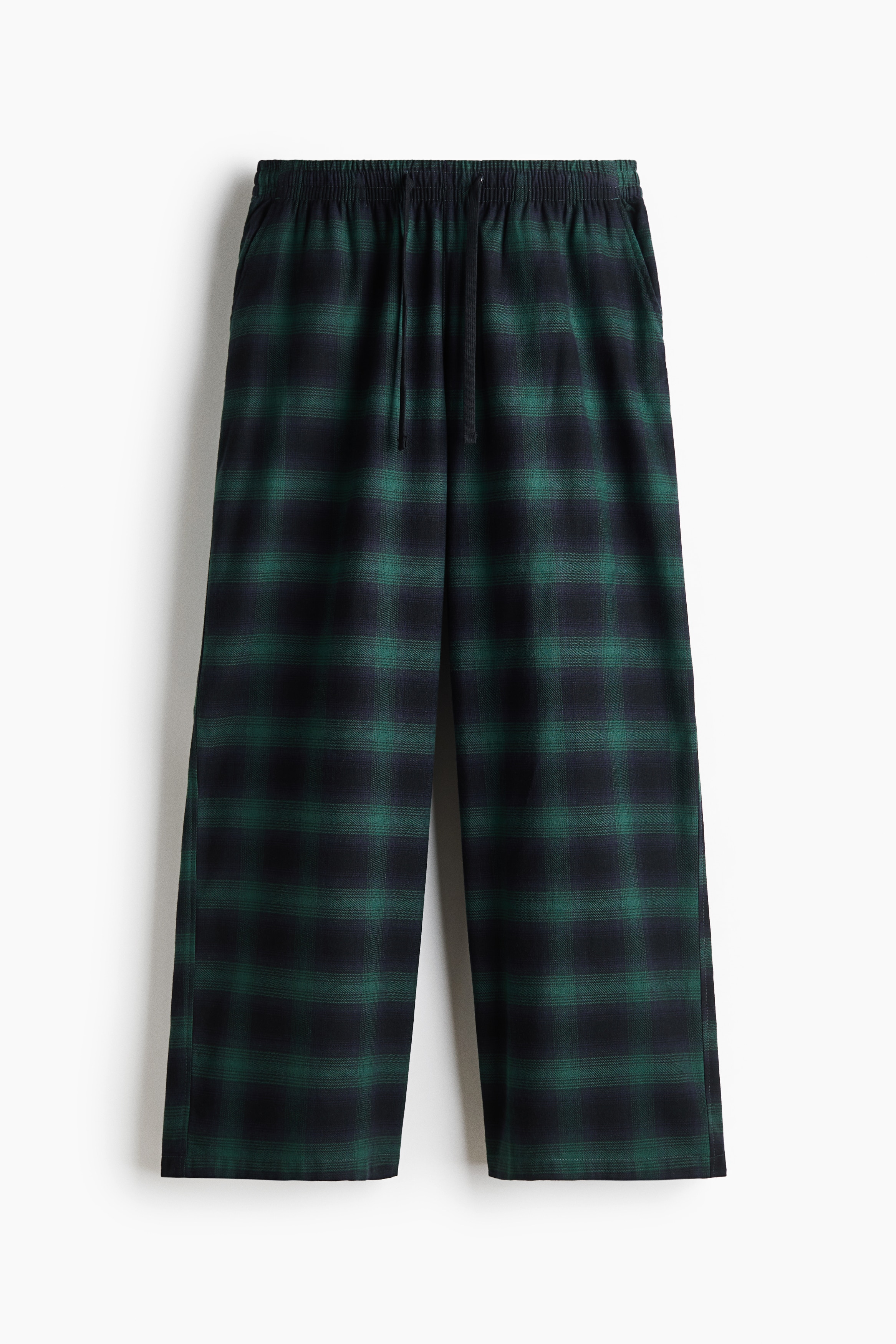 Pyjama trousers in green and black plaid cotton flannel are being worn by the model, featuring a wide-leg silhouette and an elasticated waistband with a drawstring. The trousers are styled with a light grey quarter-zip sweatshirt and grey socks.