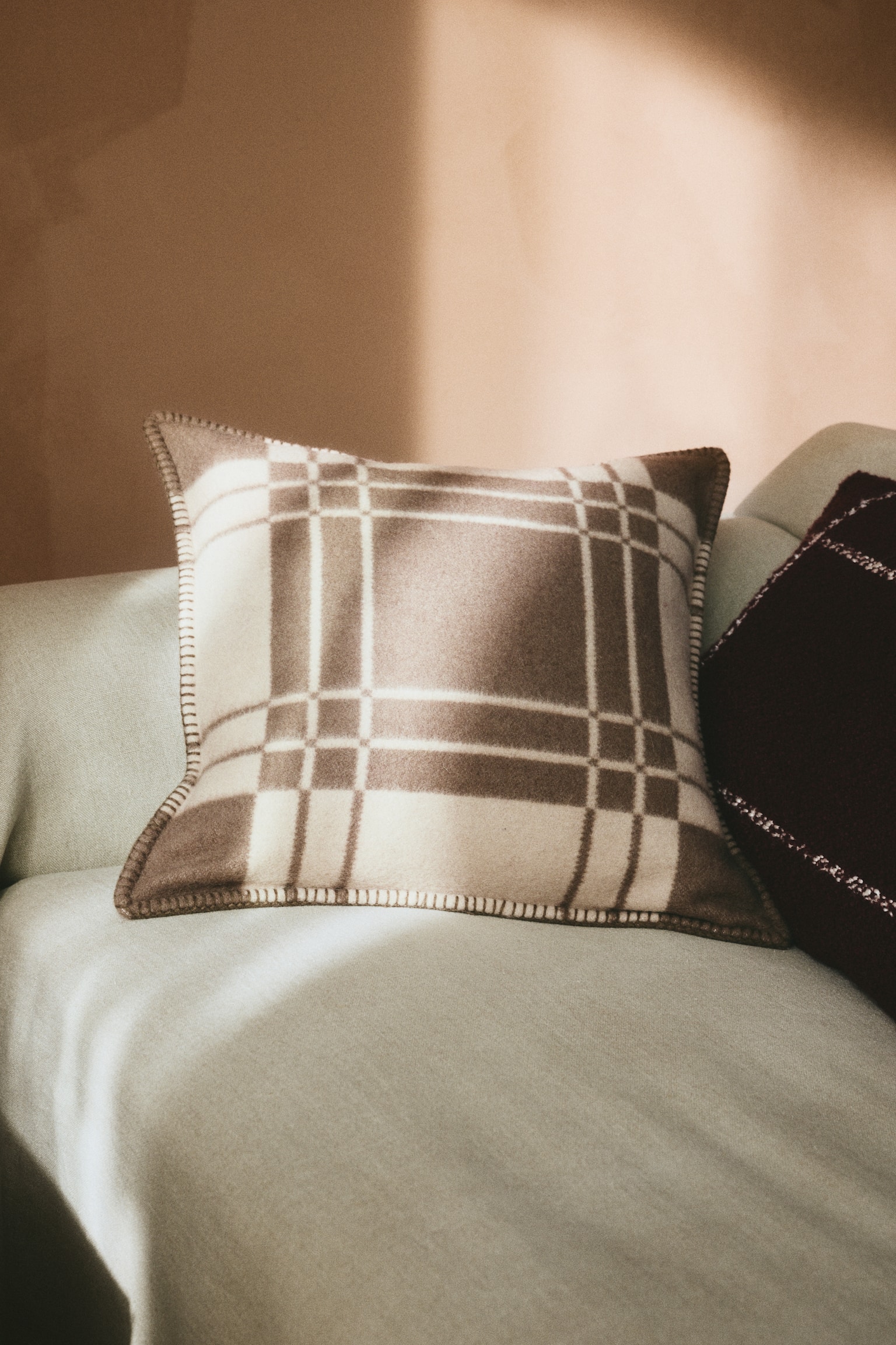 A square cushion cover is displaying a brown and cream checked pattern on its front side, featuring a soft, brushed texture. The cover is showing decorative blanket stitching along the edges while resting on a light-coloured sofa. - 1