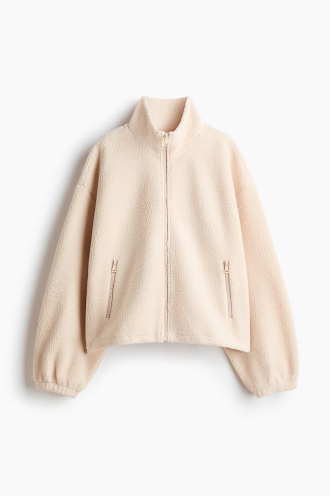 Une veste en polaire beige clair dotée d'une fermeture éclair sur toute la longueur à l'avant, d'un col montant et de deux poches zippées. Ses longues manches se terminent par des poignets froncés. - 1