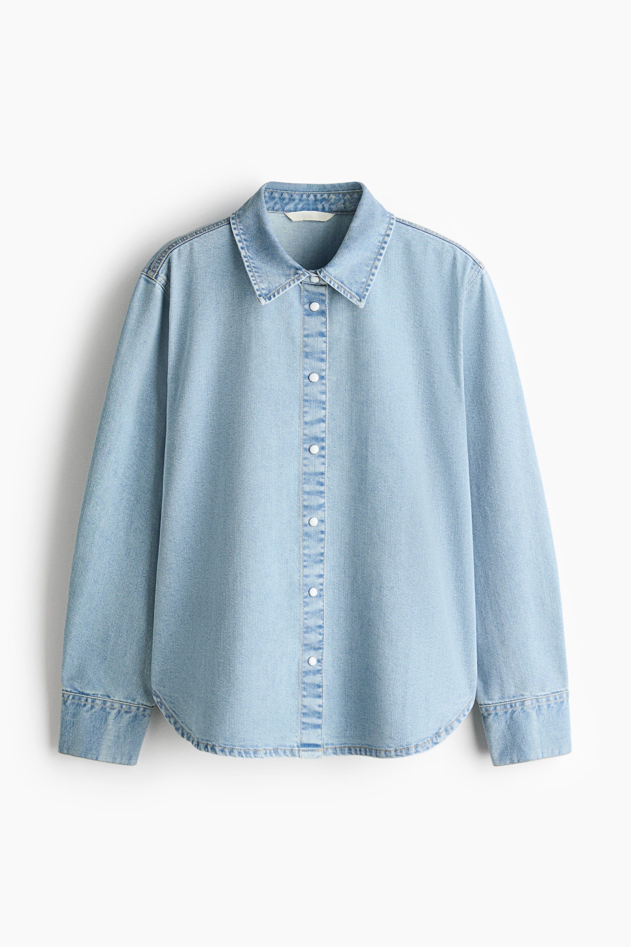 View larger image: A light blue denim shirt is front-facing, featuring a pointed collar, a full-length button placket with white buttons, long sleeves with buttoned cuffs, and a curved hem.
