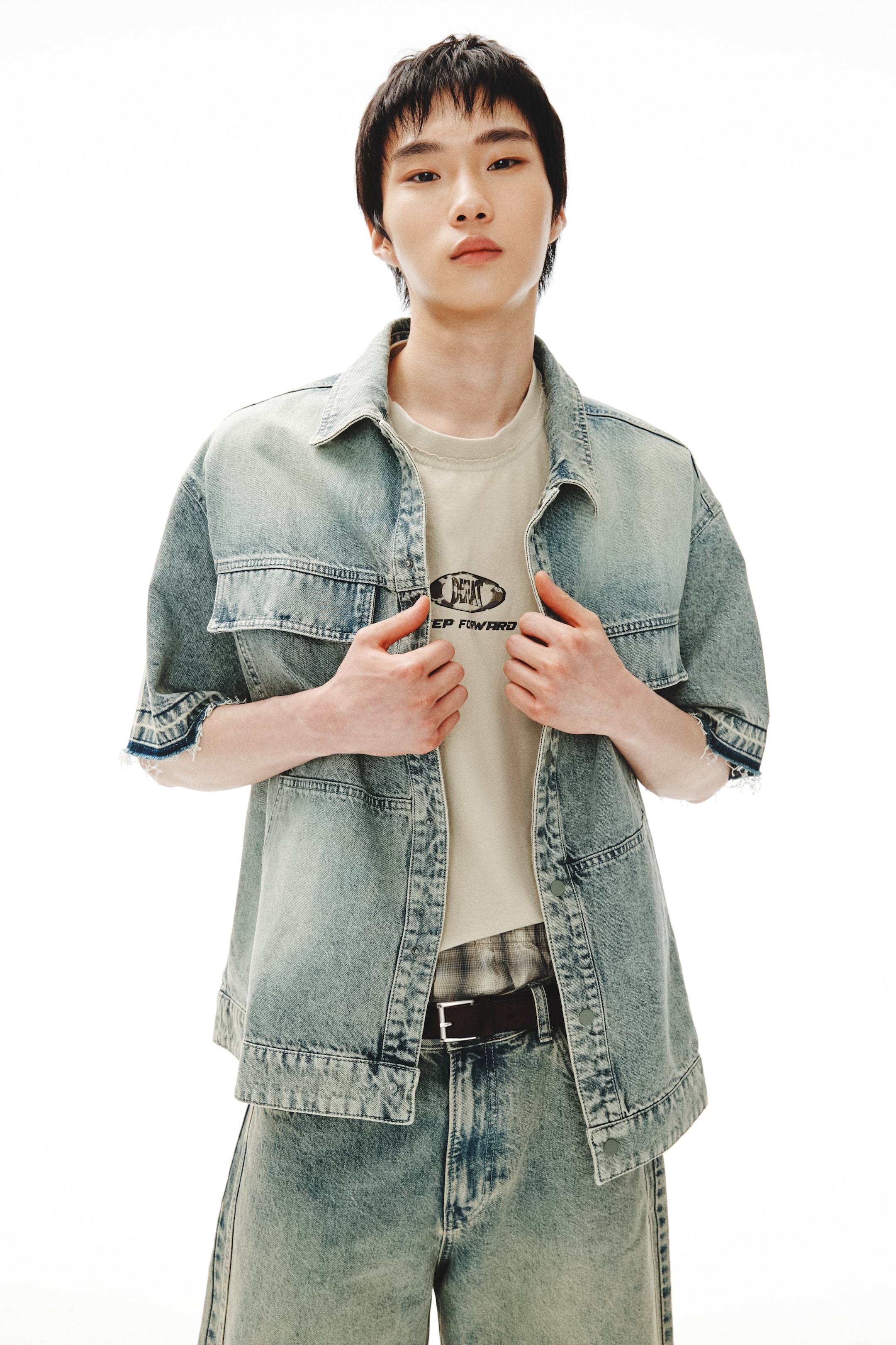 Xem ảnh lớn hơn: Một chiếc áo sơ mi denim màu xanh nhạt đã được giặt nhẹ, tay ngắn đang được mặc mở ra bên ngoài một chiếc áo thun cổ tròn màu be nhạt, có in hình "KEEP FORWARD". Chiếc áo sơ mi này được kết hợp với quần short denim màu xanh nhạt đã được giặt nhẹ phù hợp, có kiểu dáng ống rộng và một chiếc thắt lưng đen với khóa bạc.