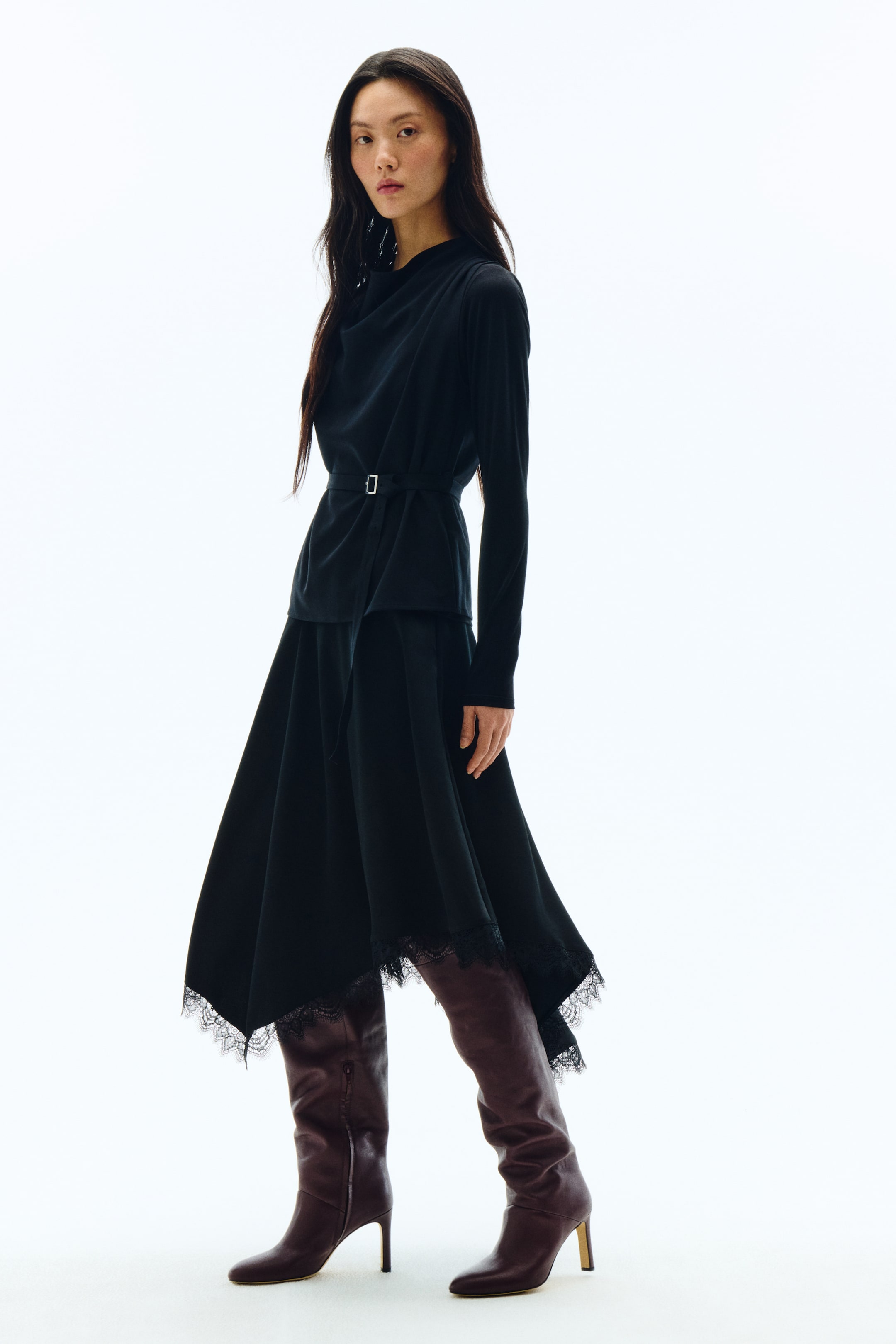 拡大画像を表示: 黒の長袖トップスにドレープの効いたカウルネックをあしらい、ウエストには長方形のシルバーバックル付きの細いブラックベルトでフィット感を持たせています。トップスはブラックのミディ丈スカートの上にレイヤードされており、スカートはアシンメトリーヘムとブラックレーストリムがデザインされています。